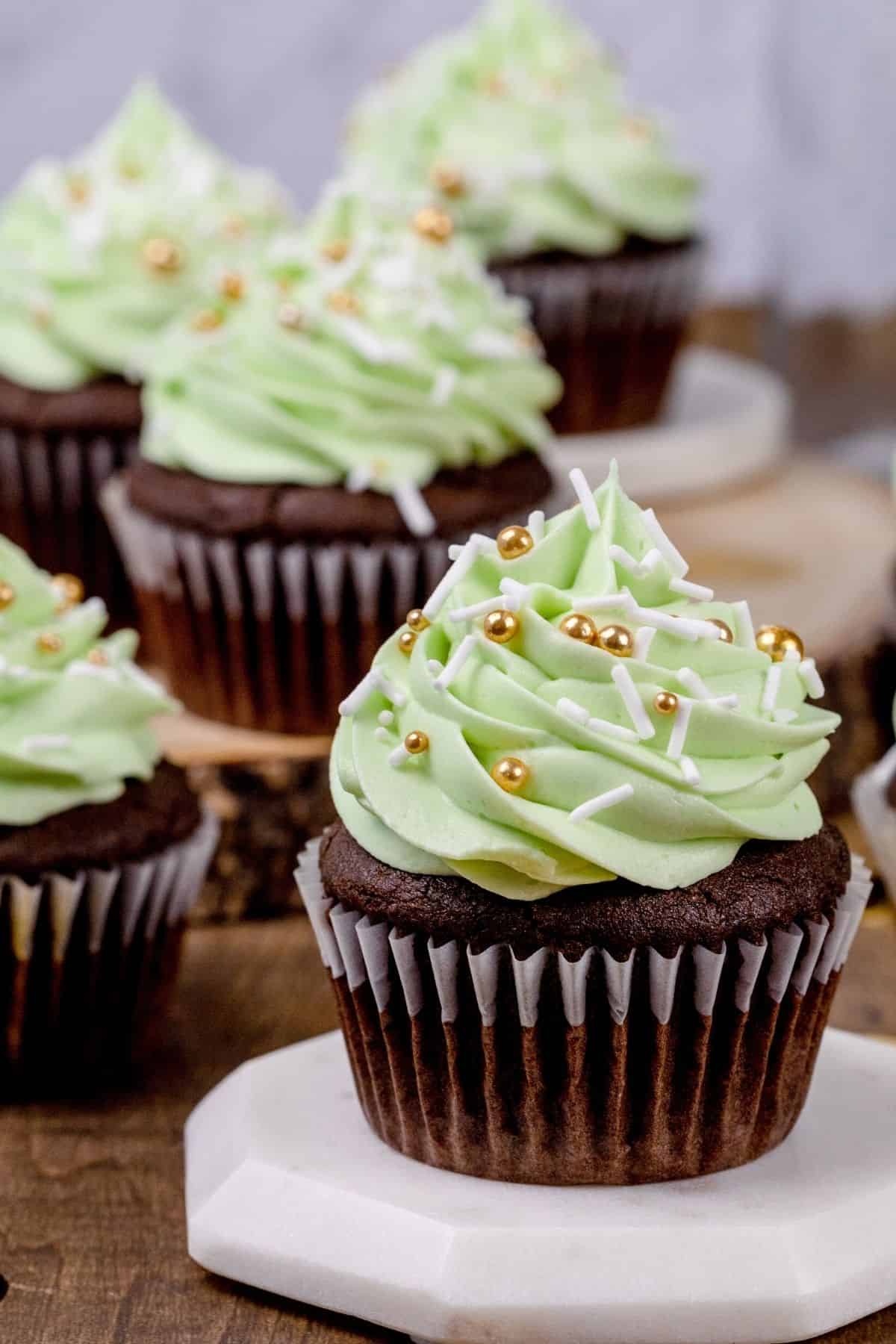 Close up of a single chocolate cupcake on a white platform. Green mint frosting is on top with gold and white sprinkles. More cupcakes are blurred in the background.