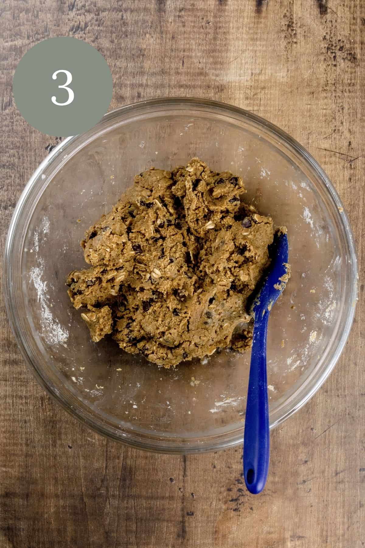 The finished cookie dough in a glass bowl with a blue spatula on a wood table top. A green circle with the number 3 is in the top left corner.