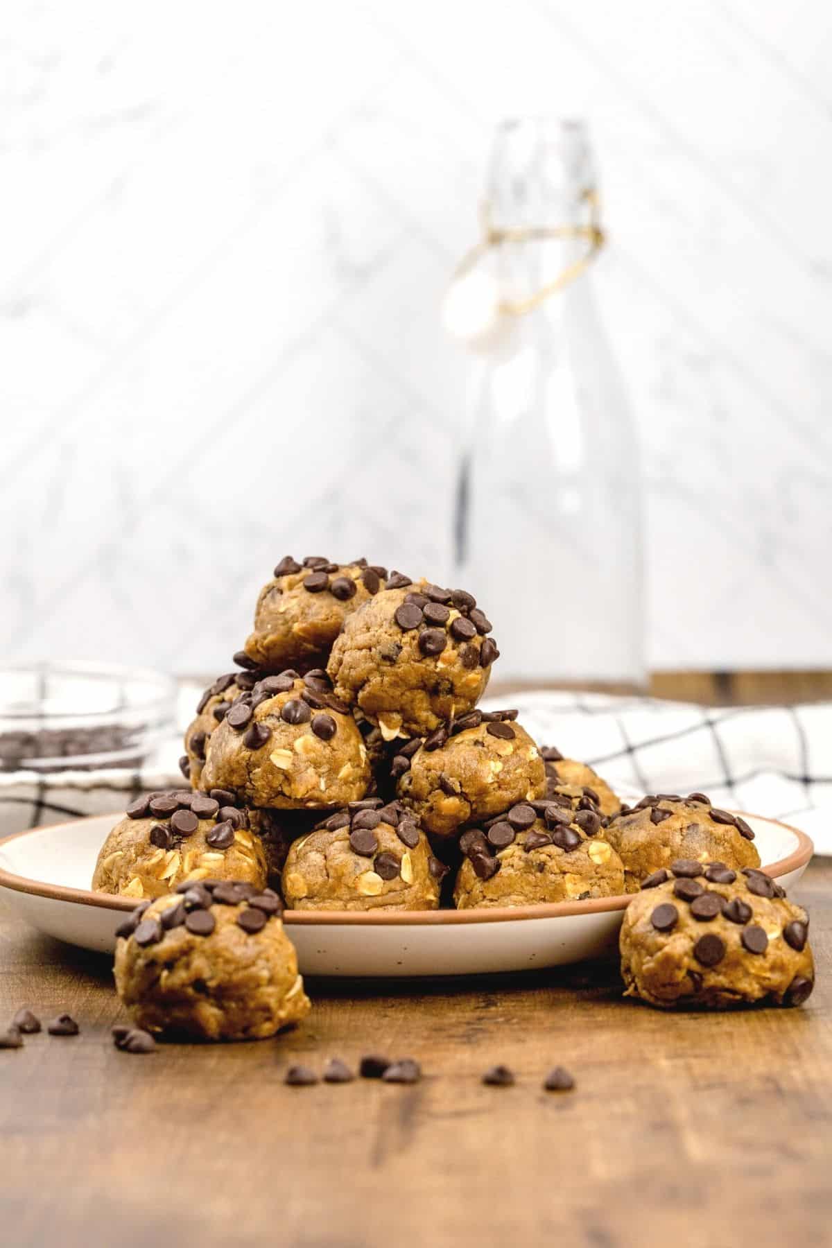 A pile of no bake chocolate chip cookie dough balls on a beige plate on a wood table in the kitchen. A towel and glass jar are blurred in the background. Mini chocolate chips are sprinkled around.