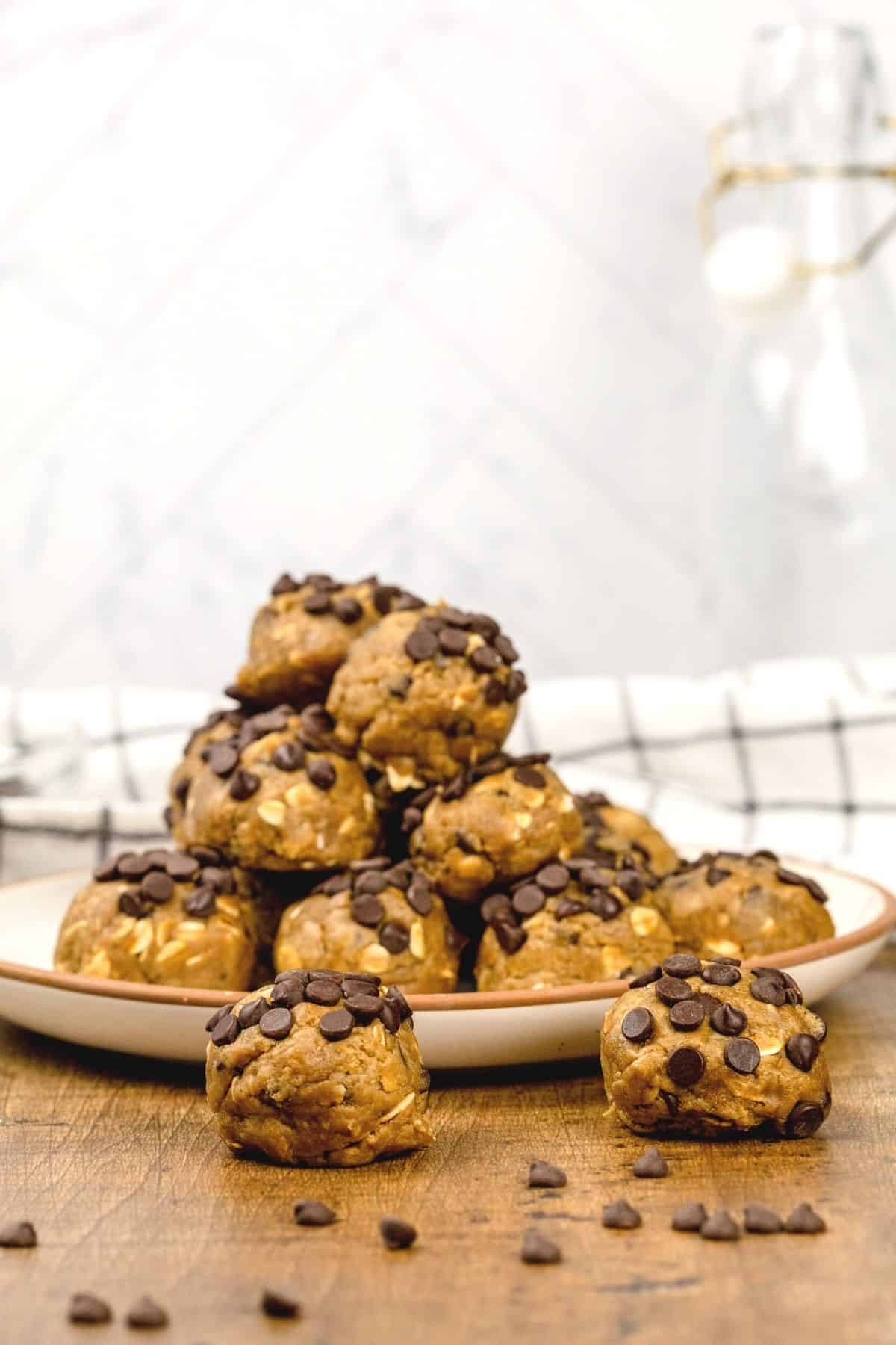 A pile of no bake chocolate chip cookie dough balls on a beige plate on a wood table in the kitchen. A towel and glass jar are blurred in the background. Mini chocolate chips are sprinkled around. The focus in on the two dough balls in front of the plate.
