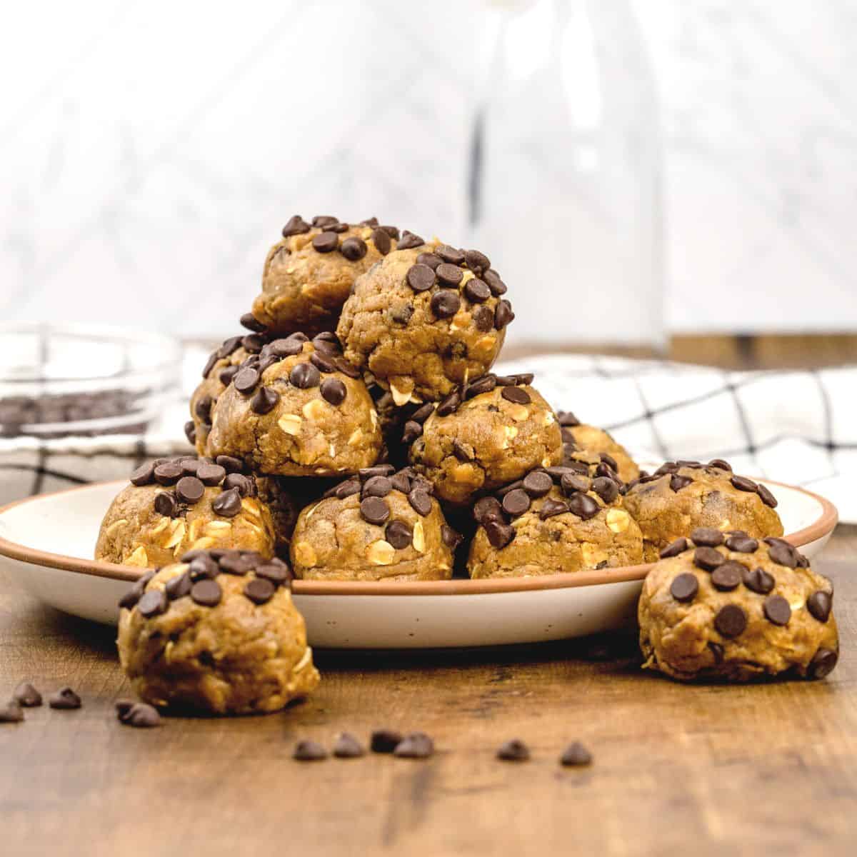 A square cropped image of multiple no bake chocolate chip cookie dough balls on a beige plate on a wood table in the kitchen. A towel and glass jar are blurred in the background. More mini chocolate chips are scattered around.