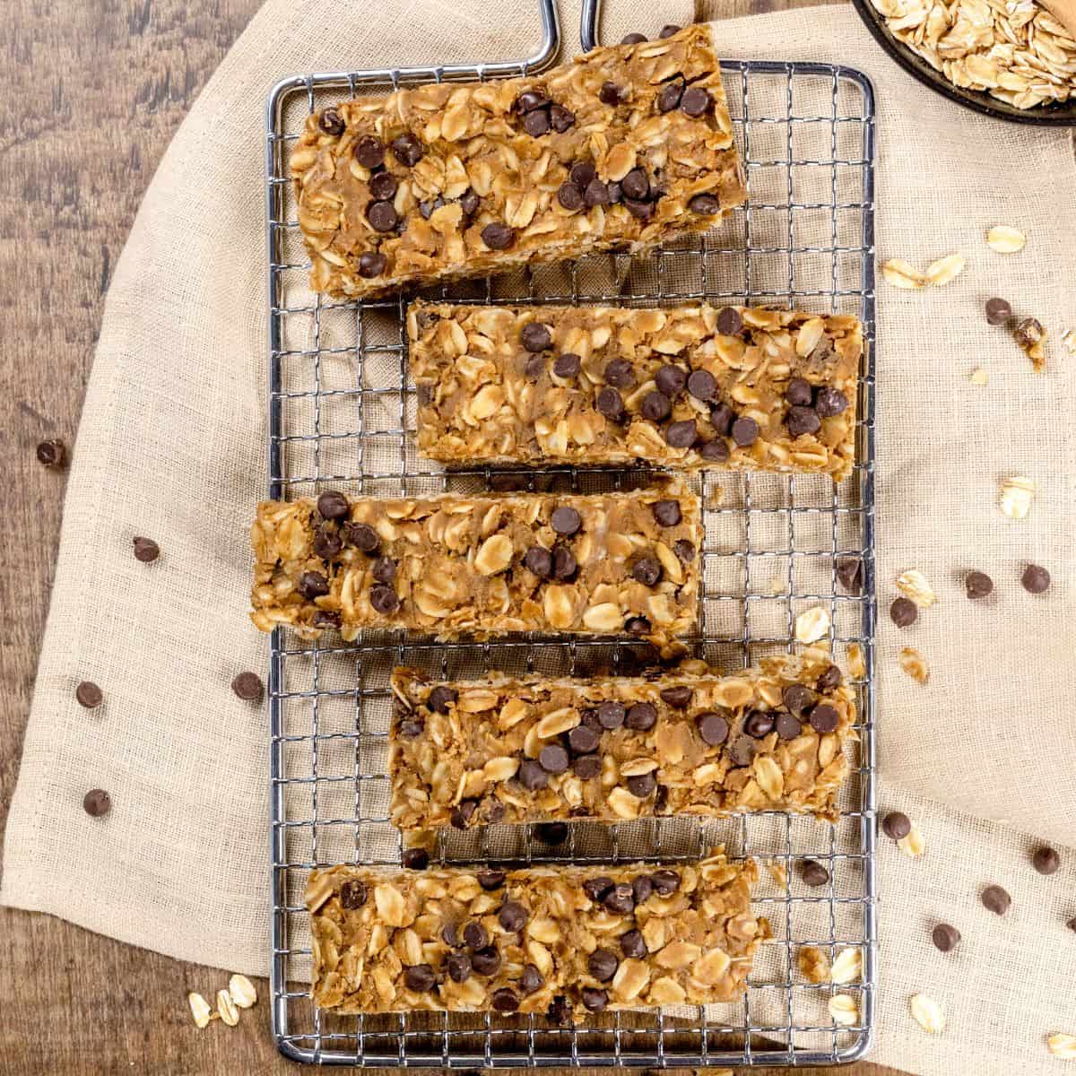 5 nut free granola bars on a metal cooling rack on a wood table. A soft linen is under the granola bars. Toasted oats and mini chocolate chips are sprinkled around.