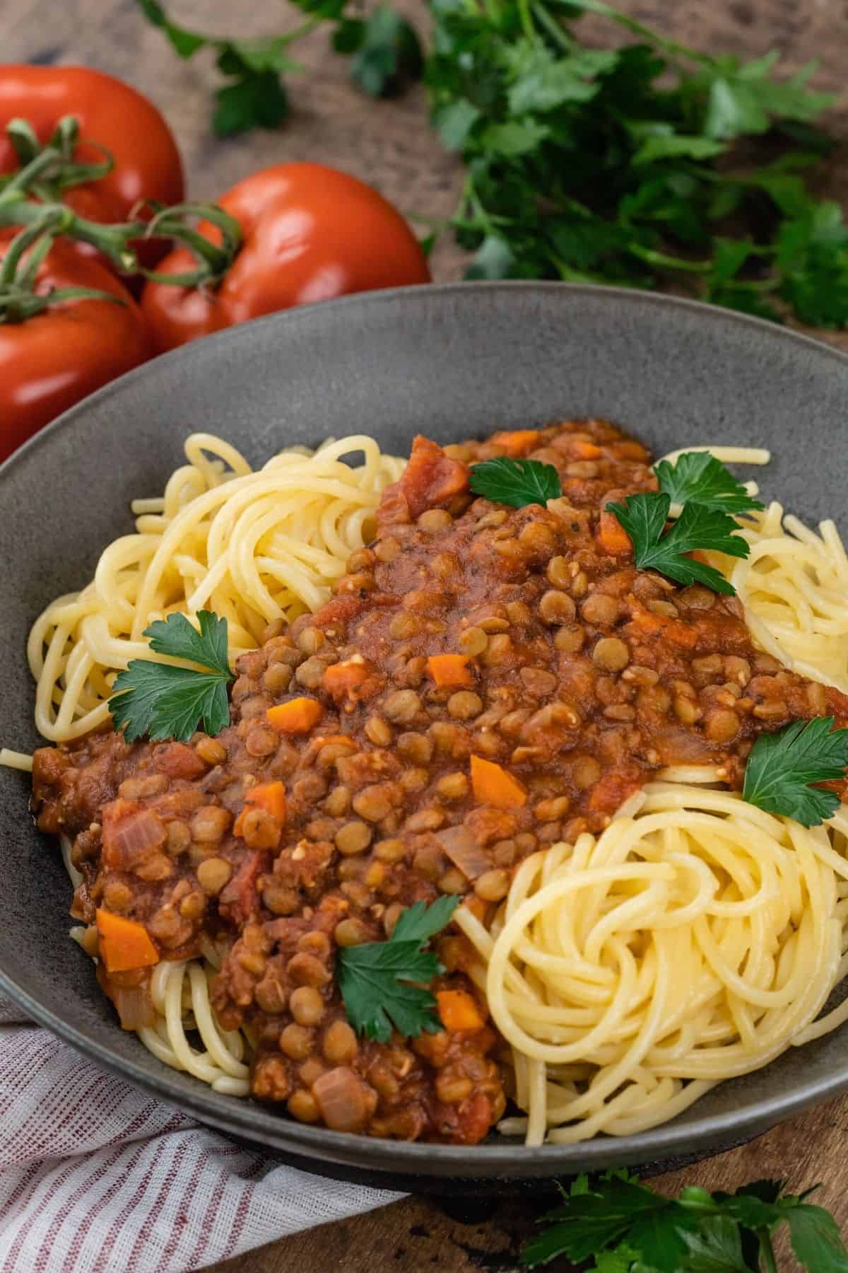 Close up of a bowl of lentil bolognese on top of spaghetti noodles. Fresh parsley and tomatoes are blurred in the background.