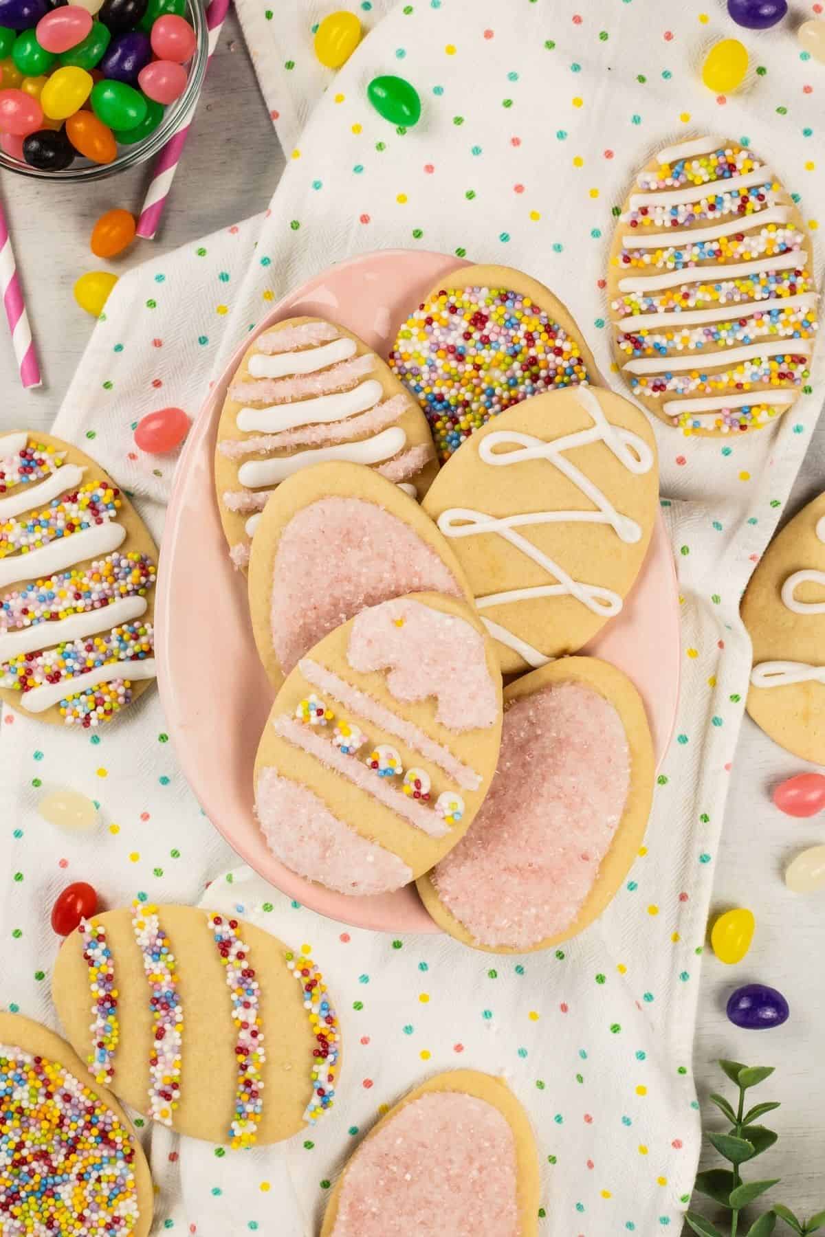 A table scape of sugar cookies for Easter cut into egg shapes. A multicolored towel and lots of jelly beans are also on the table.