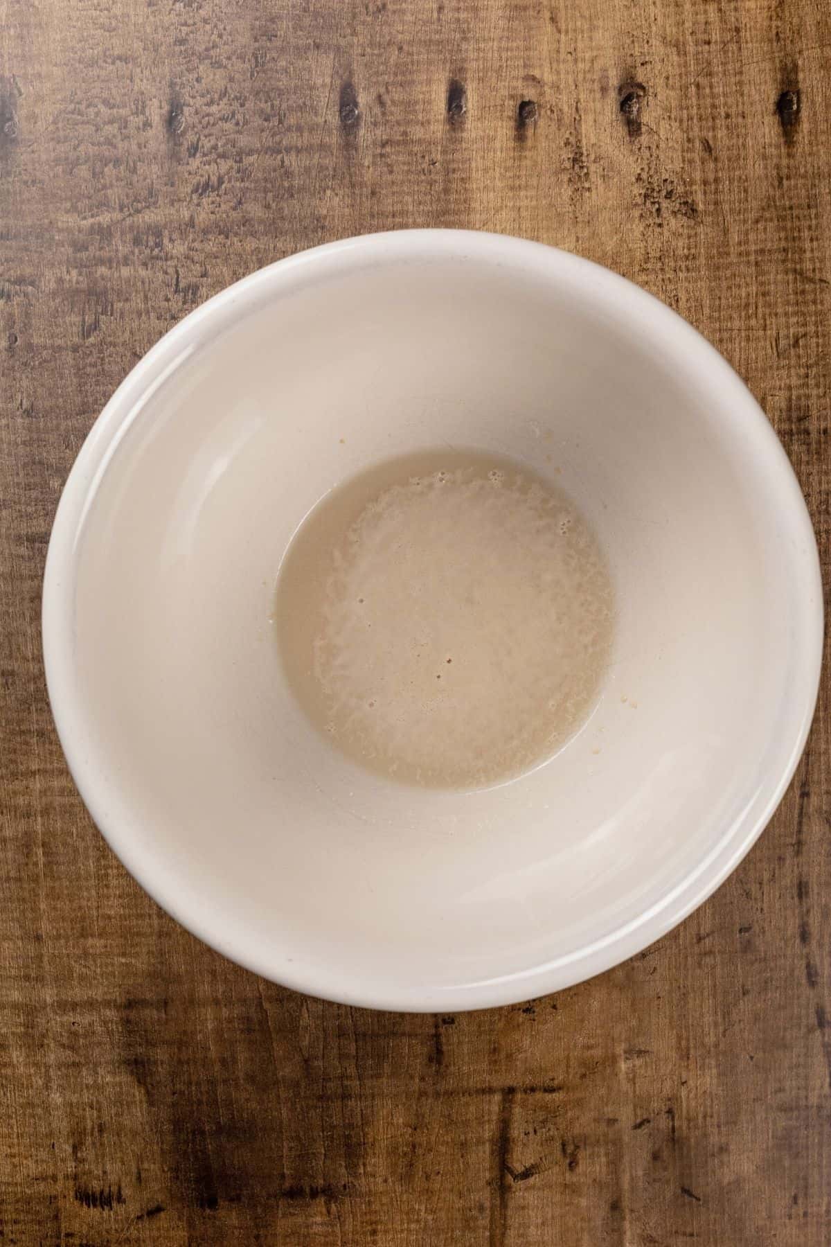 A ceramic bowl is filled with yeast and warm water. It rests on a wood tabletop.