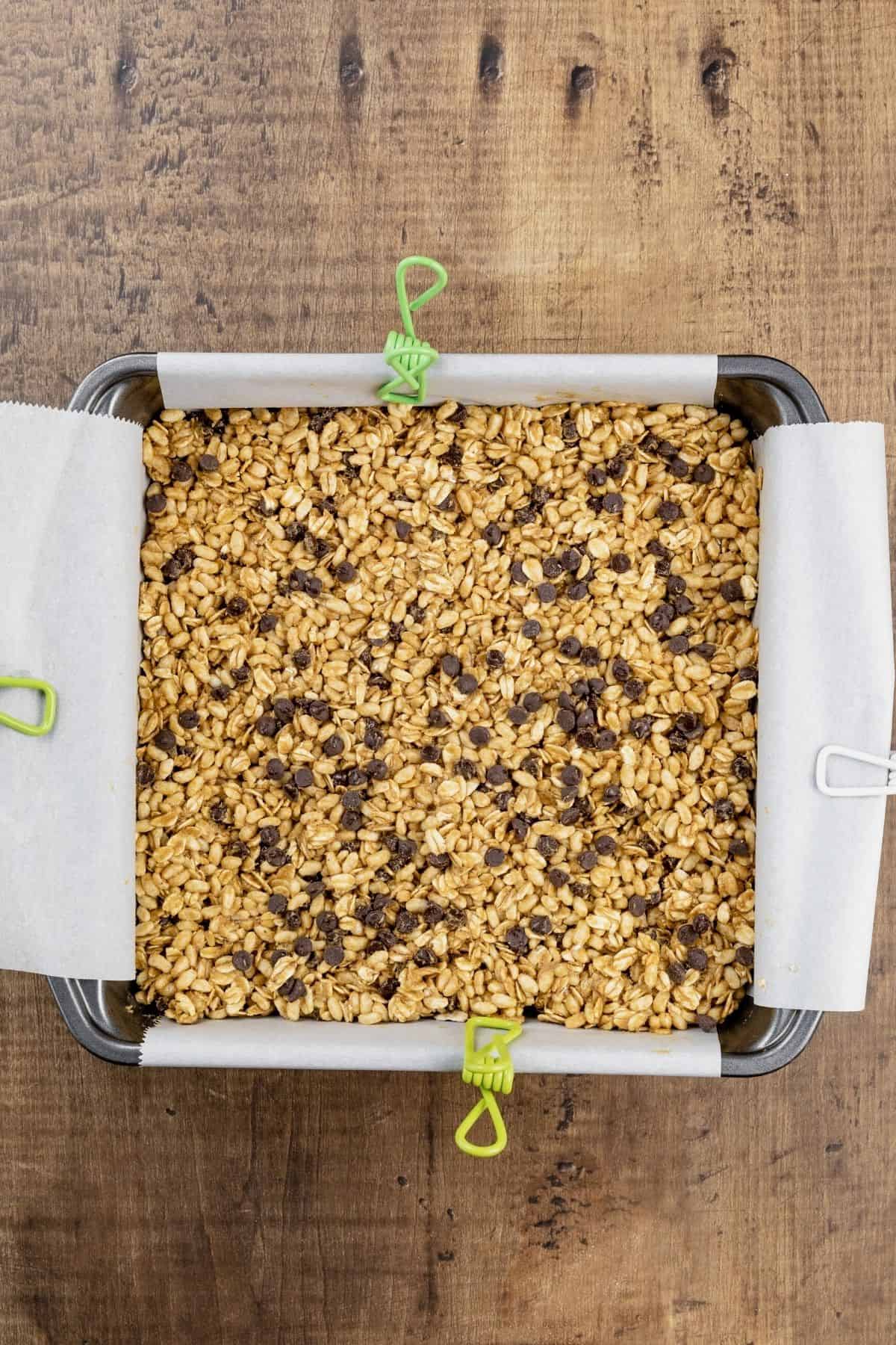 A metal 8x8 pan is lined with parchment paper and filled with the granola bar mixture that has been pressed into place. It rests on a wood table.