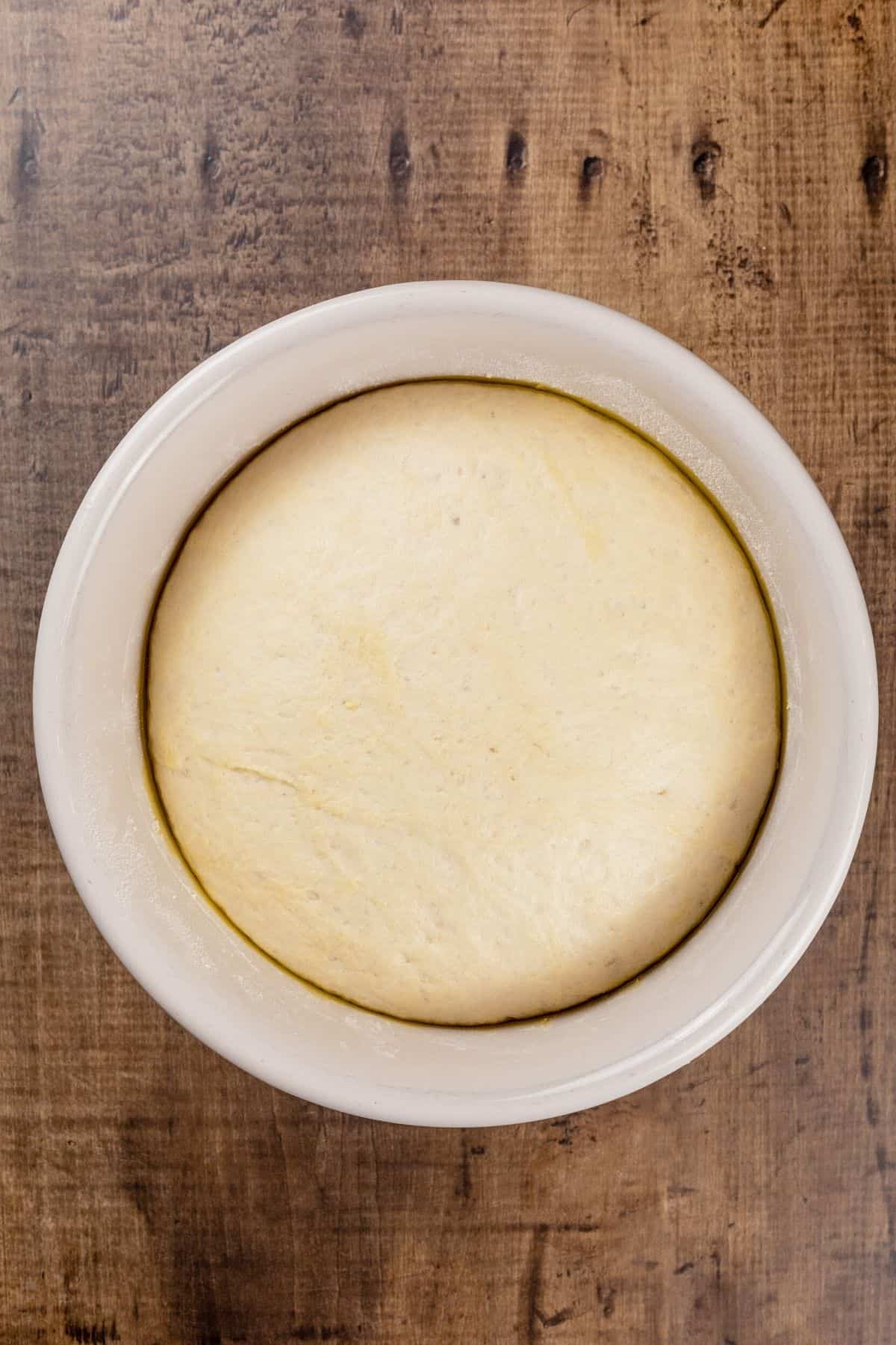 The fully risen pizza dough in the same ceramic bowl. You can see how full the bowl has become. It is resting on a wood tabletop.