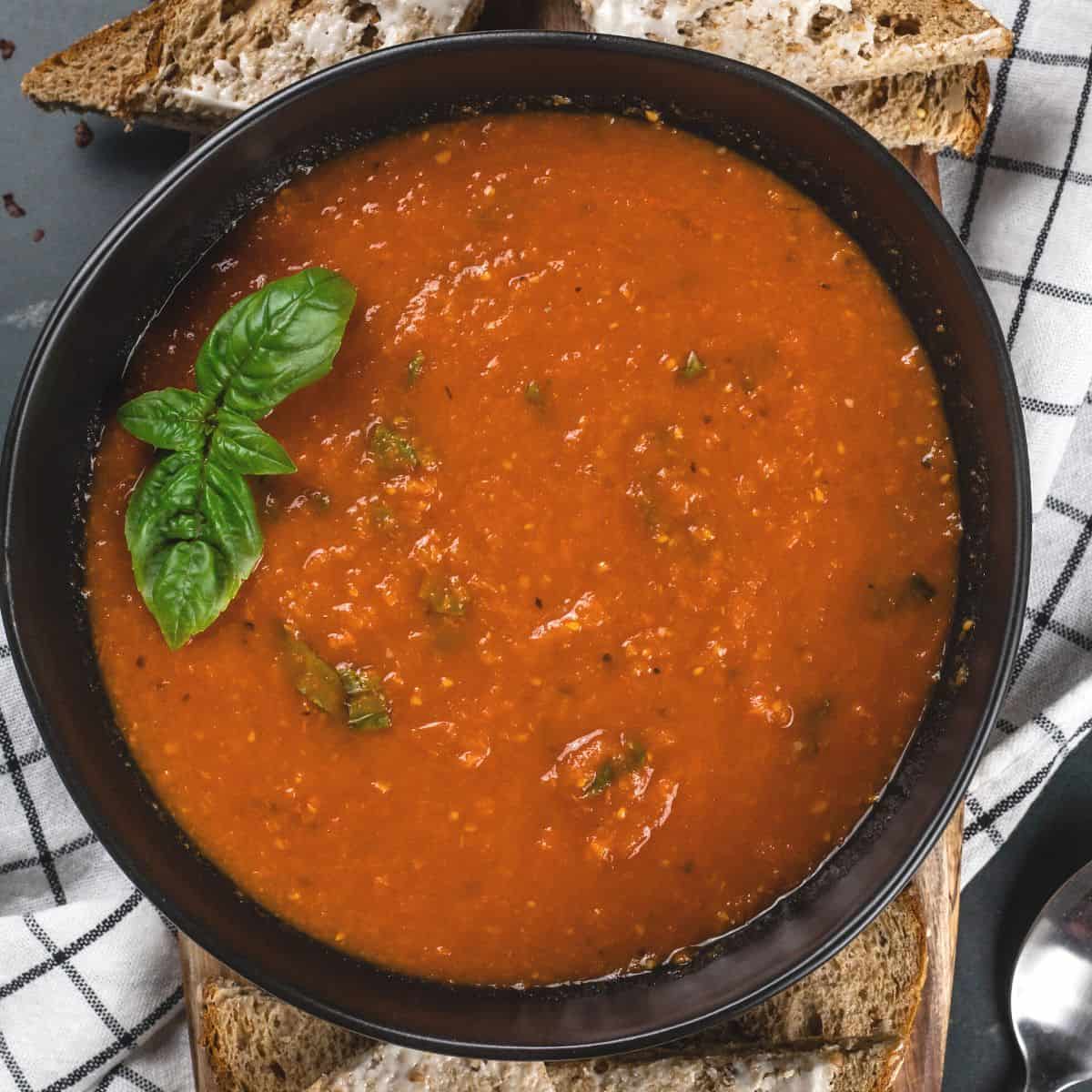 A big black bowl filled with roasted tomato basil soup. Fresh basil is sitting on top of the soup. A white and blue towel is under the bowl. Slices of hearty bread and butter are seen.