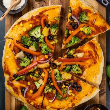 Vegan pizza on a wood cutting board with lots of veggies on top like red pepper and broccoli that has been cut into 6 slices.