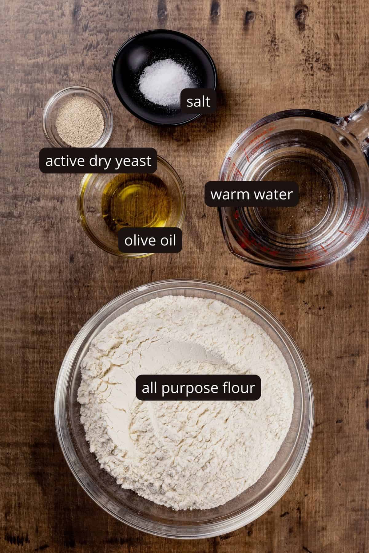 Ingredients for vegan pizza dough in various glass bowls on a wood tabletop. Black and white labels have been added to name each ingredient.