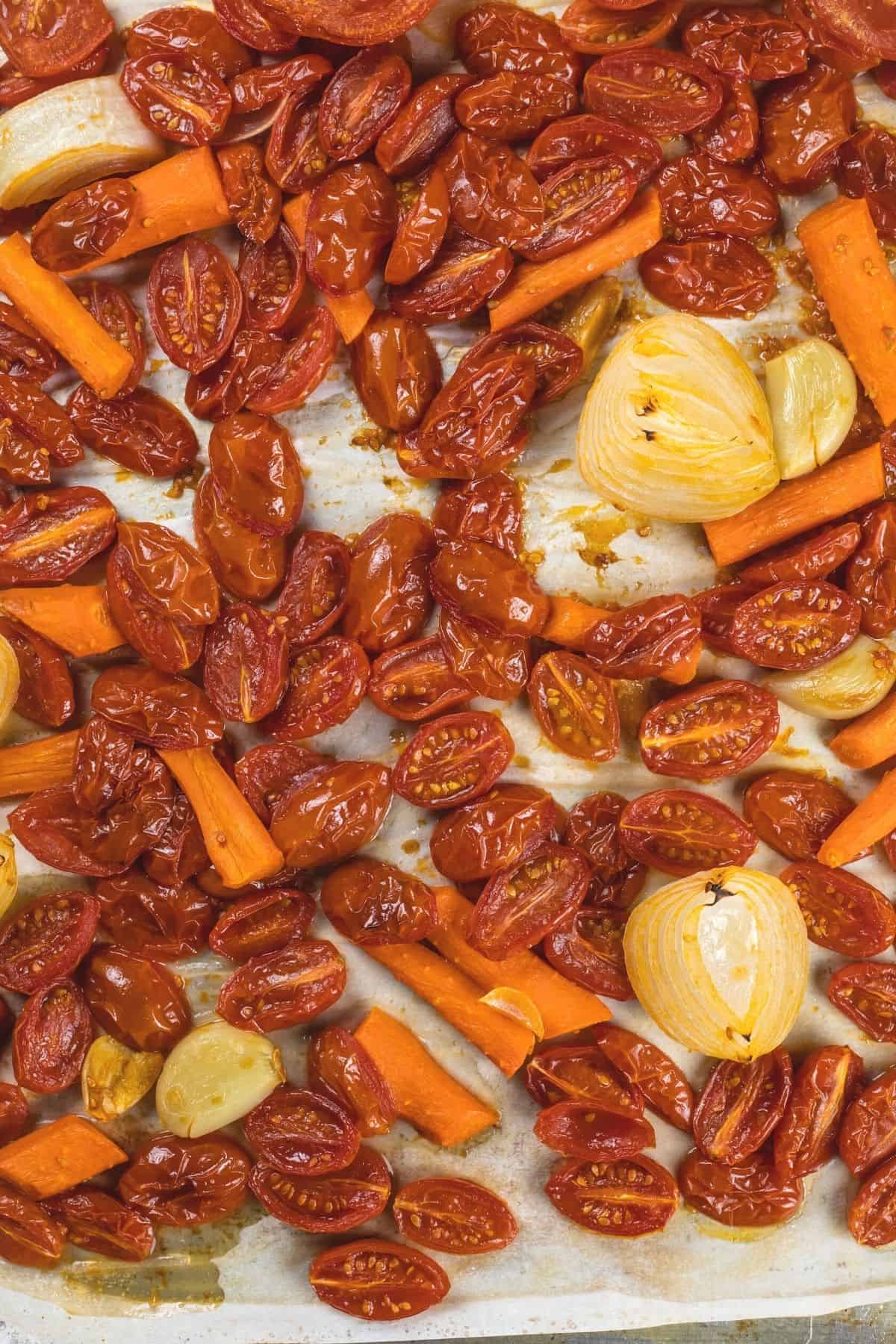 Roasted vegetables like tomatoes, onions, garlic, and carrots on a silver baking tray with parchment paper.