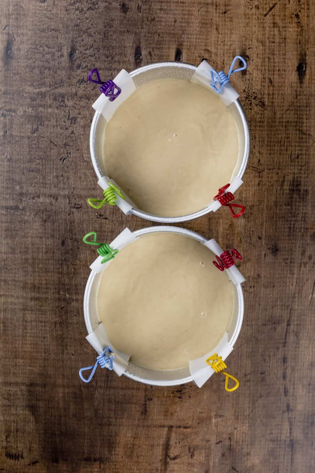 Two cake pans are filled with the finished cake batter. Strips of parchment paper are held in place by small clips.