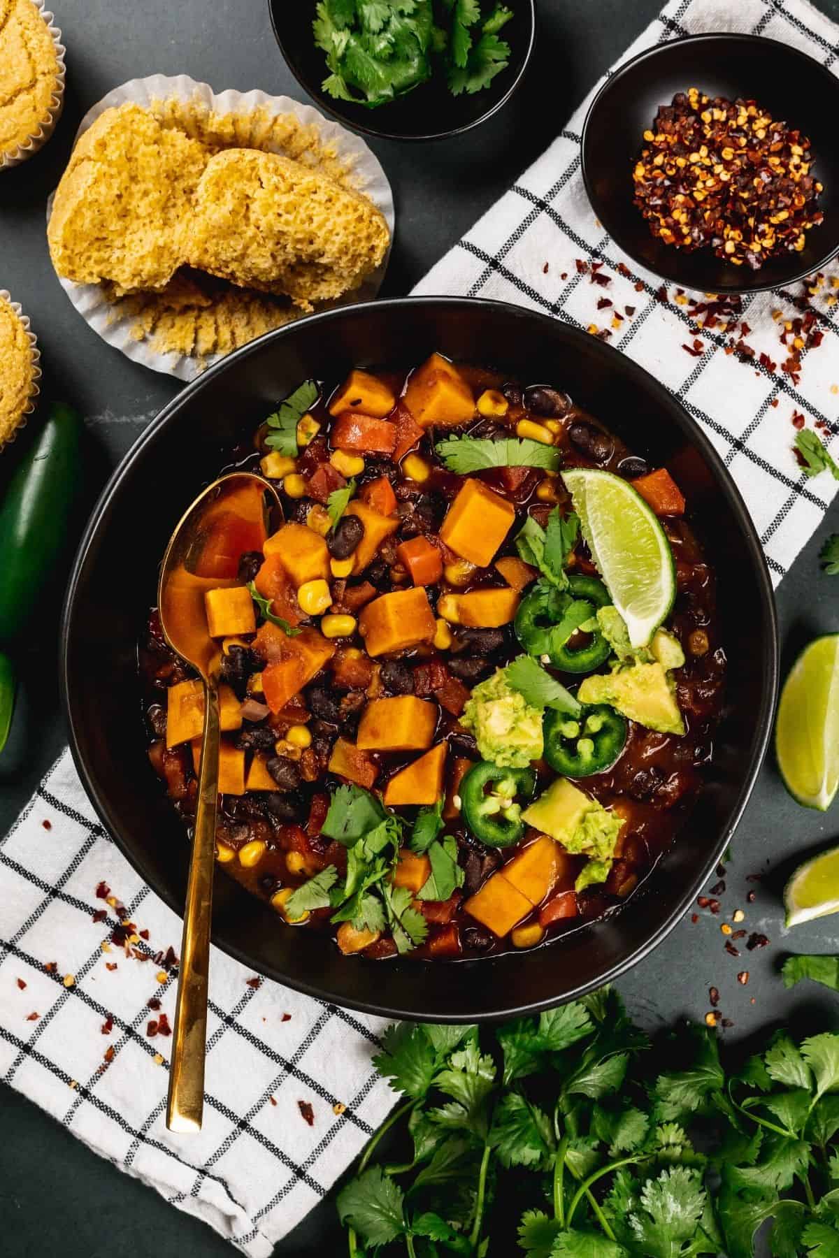 A big black bowl filled with sweet potato and black bean chili with many toppings on it like avocado, peppers, and cilantro. More toppings surround the bowl as it rests on a white towel.