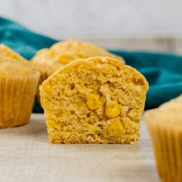 An egg free cornbread muffin is sliced in half so you can see the texture and corn kernels on the inside. It is one a white kitchen table next to more muffins. A teal towel is blurred in the background.