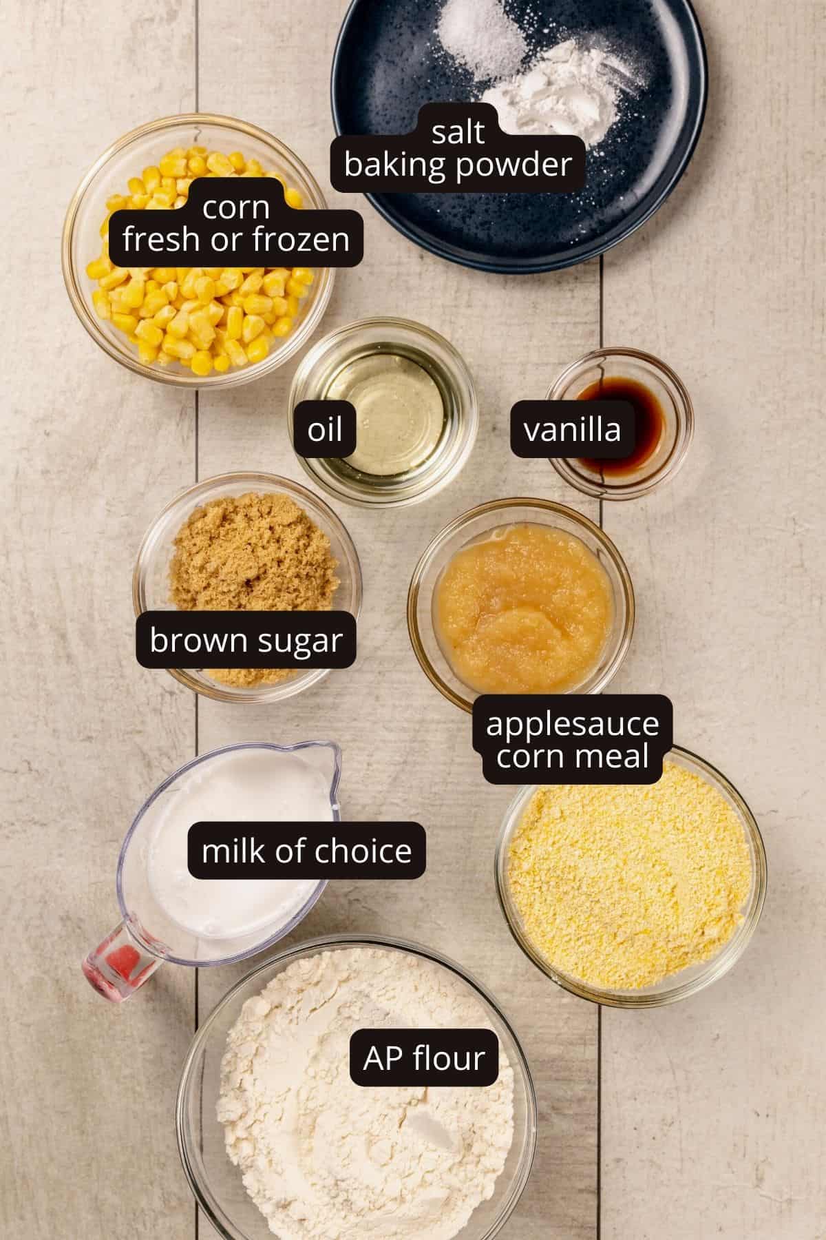 Ingredients for cornbread muffins in various glass bowls on a white wood kitchen table. Black and white labels have been added to name each ingredient.