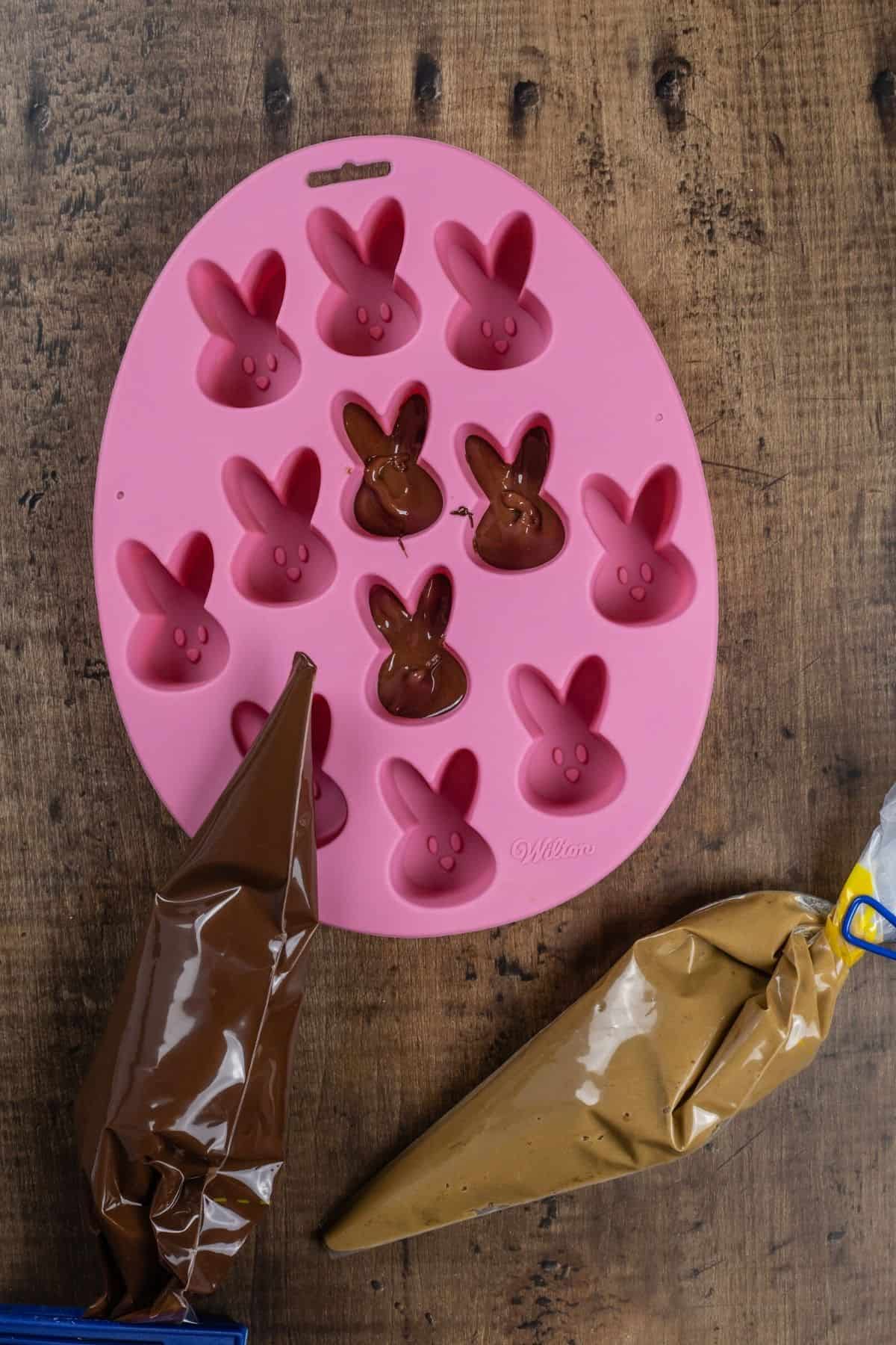 A pink bunny candy mold is being filled with a first layer of chocolate. It rests on a wood table. 2 piping bags are next to it, one filled with chocolate and the other with the sunbutter filling.