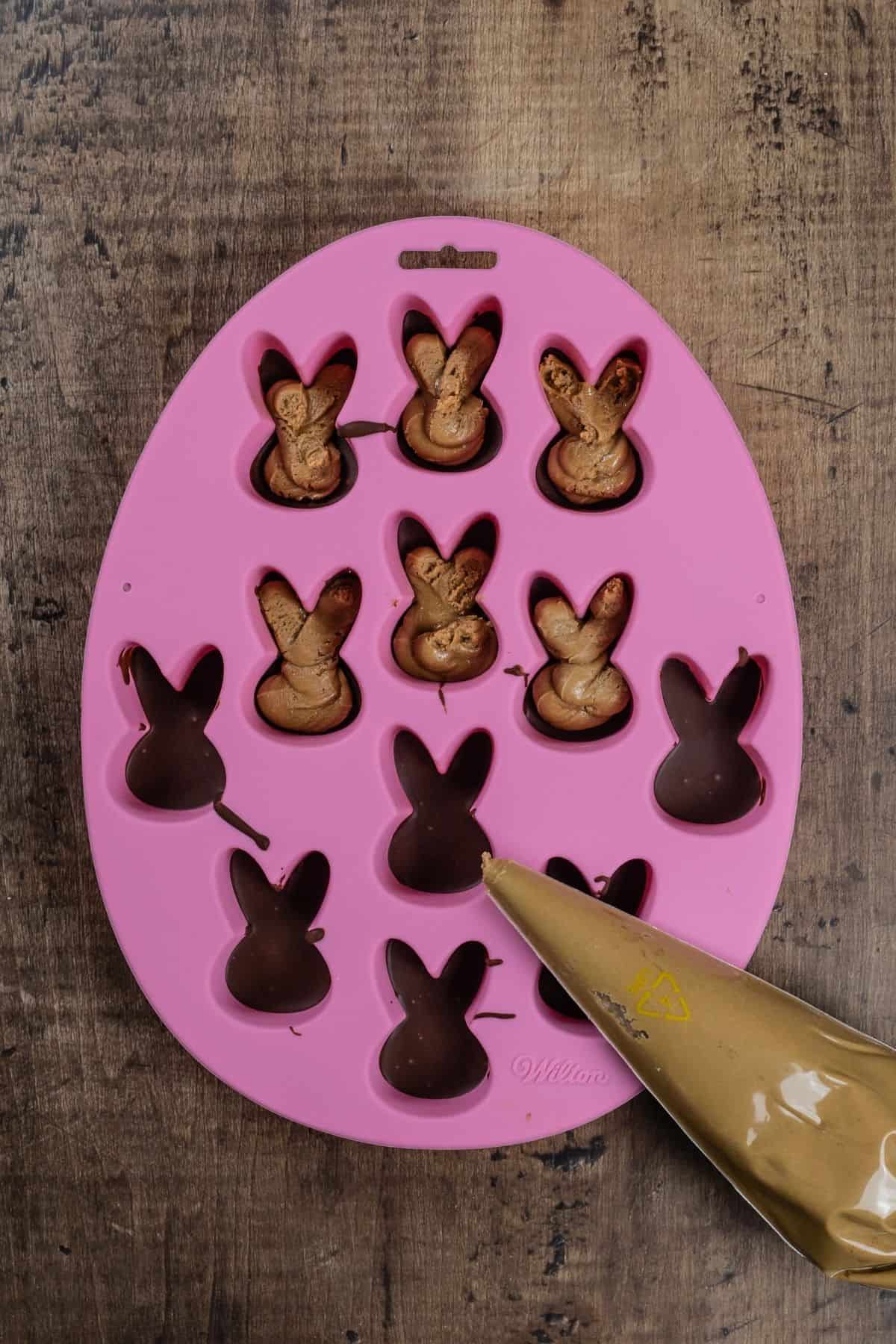 A pink bunny candy mold on a wood tabletop is being filled with sunbutter filling. A piping bag is next to the mold and is filled with the filling.