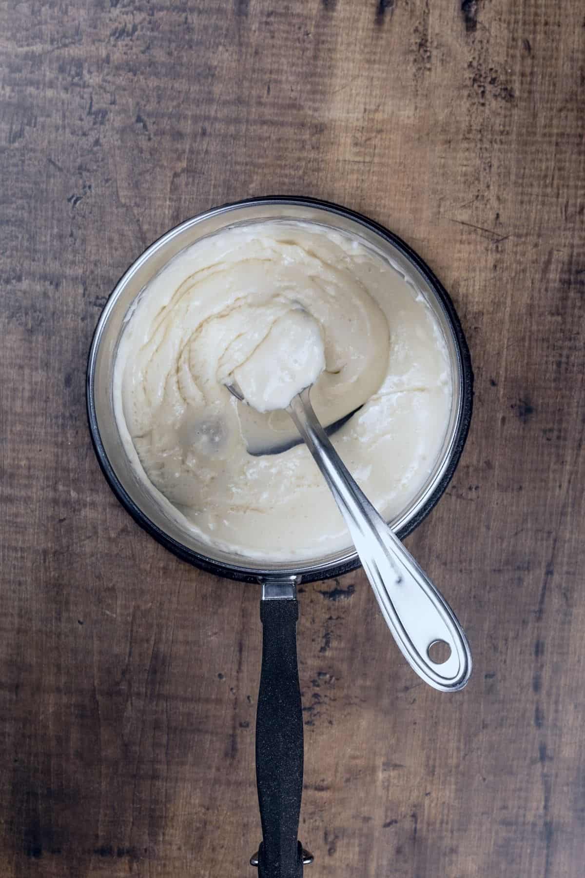 A cooking pot filled with the finished marshmallow frosting. A metal silver spoon rests in the frosting. The pot is on a kitchen table.