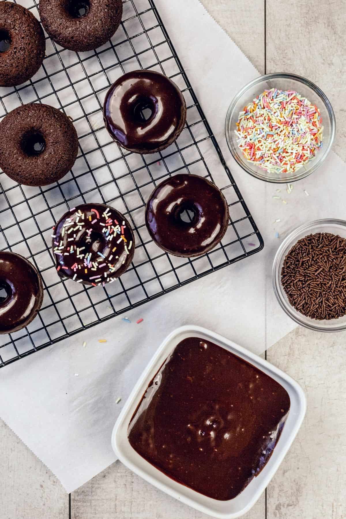 Many fully cooked chocolate donuts on a wire cooling rack next to a bowl of chocolate ganache and two small bowls filled with sprinkles. Each donut is getting dipped and sprinkled.