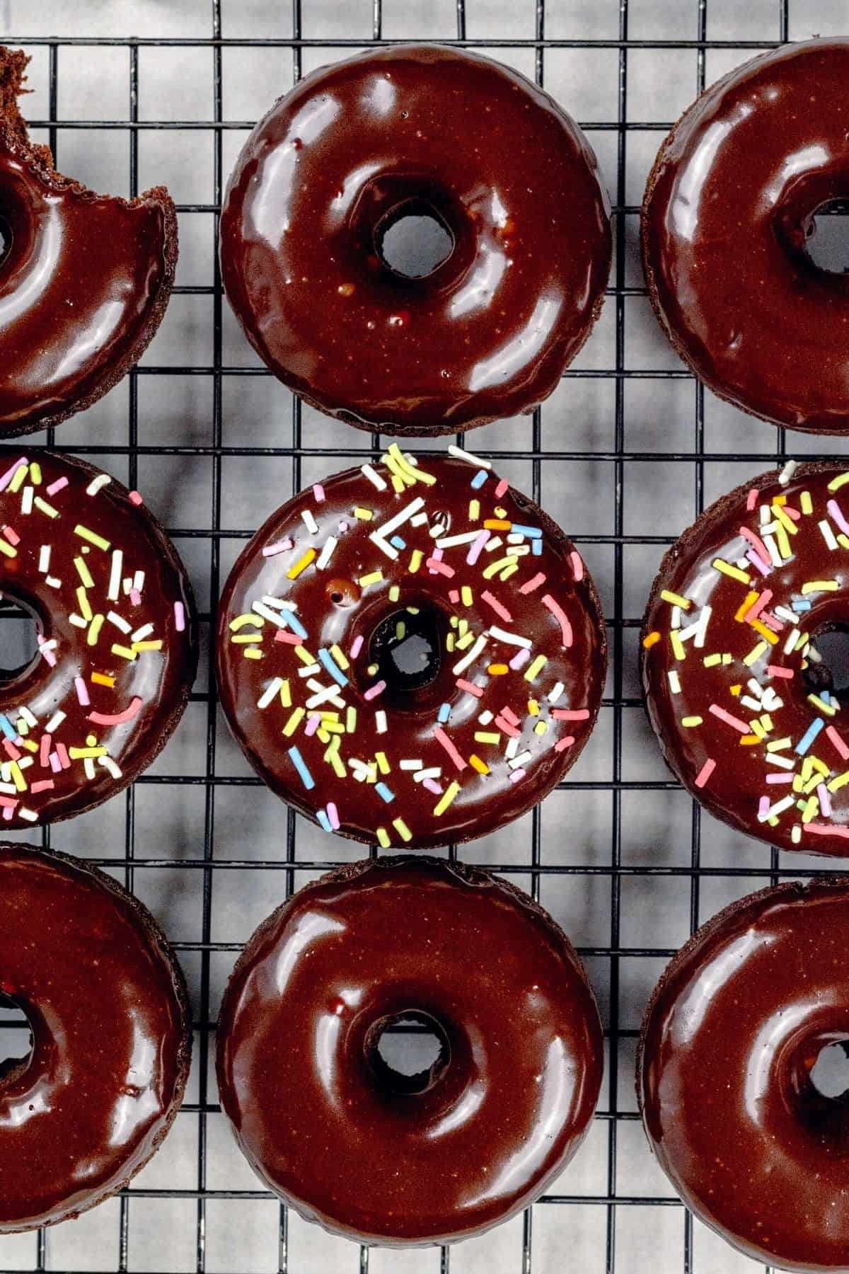 Close up of 9 frosted chocolate donuts on a wire cooling rack. 3 of the donuts have multicolored sprinkles. The top left donut has a bite taken out of it.