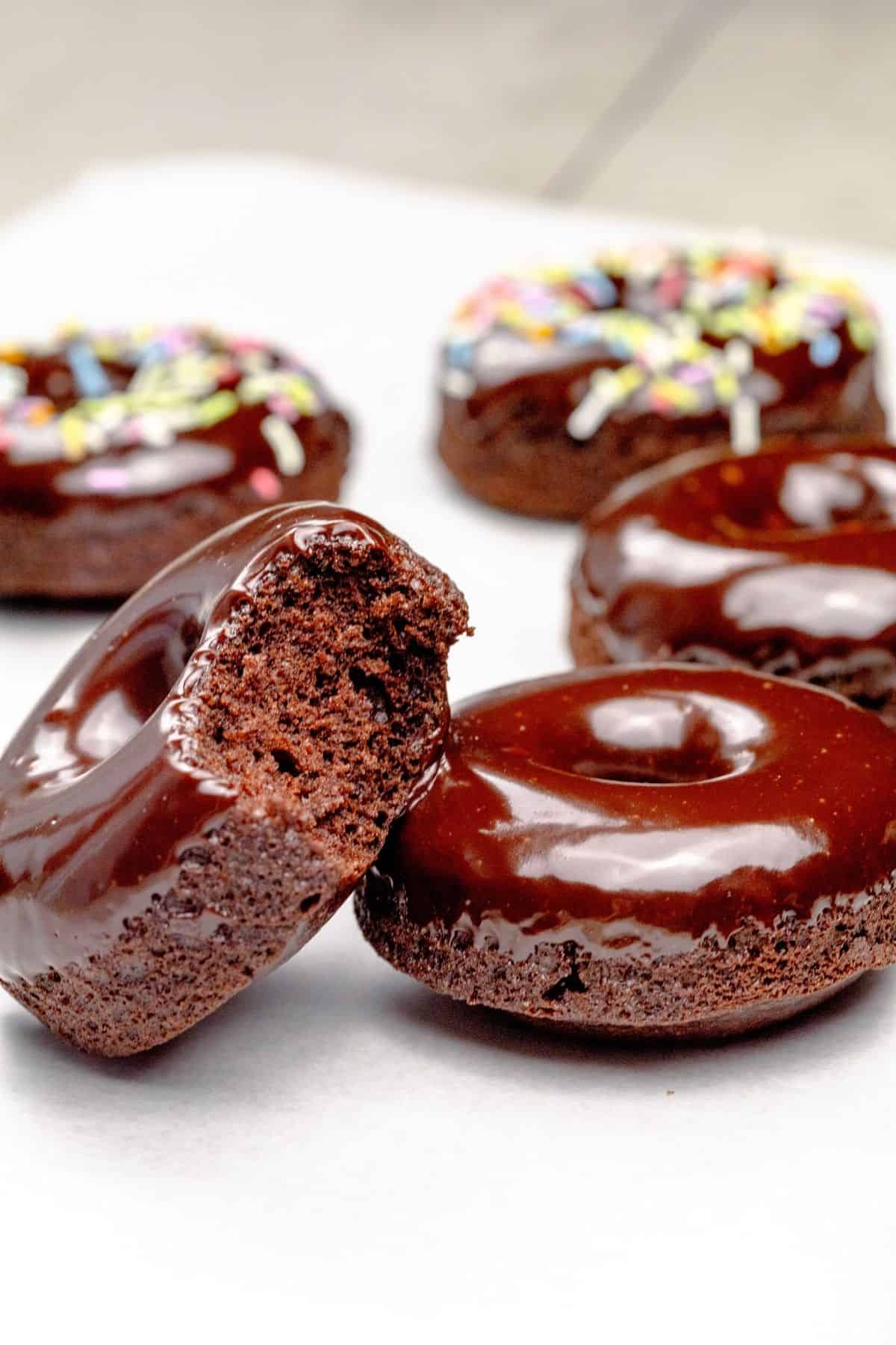2 gluten free vegan chocolate donuts are next to each other on a white parchment paper. One of the donuts is leaned against the other with a bite taken out of it. Donuts covered in sprinkles are blurred in the background.