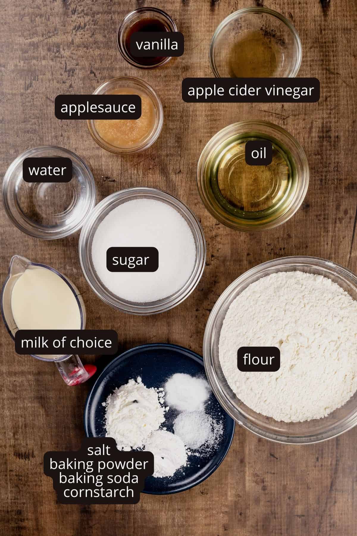 Ingredients for a vegan vanilla cake in various glass bowls are on a wood table top. Black and white labels have been added to name each ingredient.