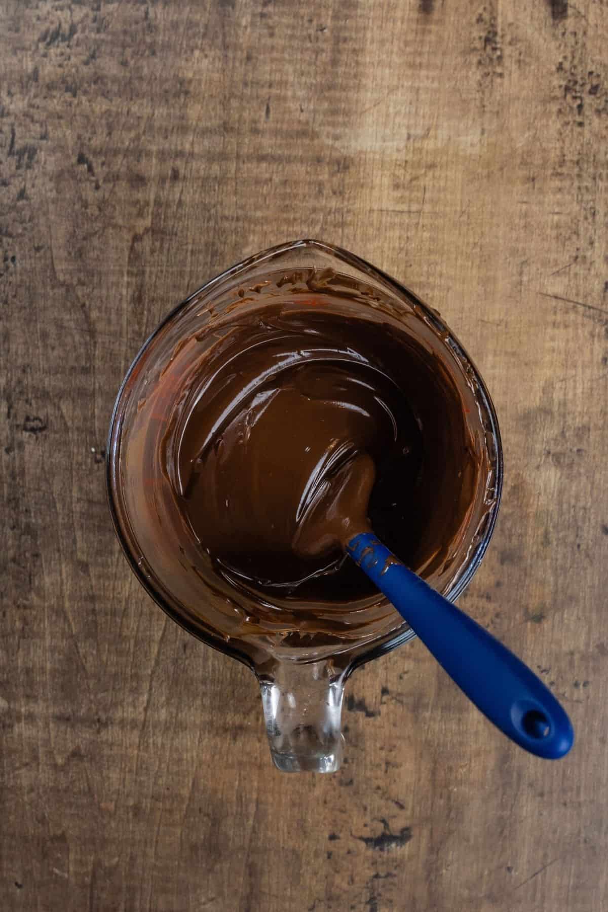 A glass measuring cup is filled with melted chocolate. A blue spatula rests in the chocolate. The cup sits on a wood tabletop.