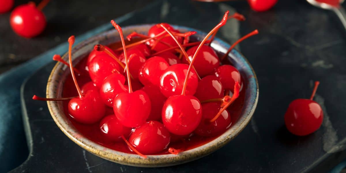 Many maraschino cherries in a dark bowl on a dark tabletop. A few cherries are scattered around the bowl.