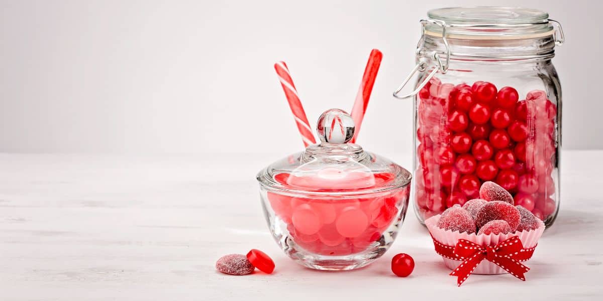 Glass jars are filled with different types of red candies. They are on a white table and background.