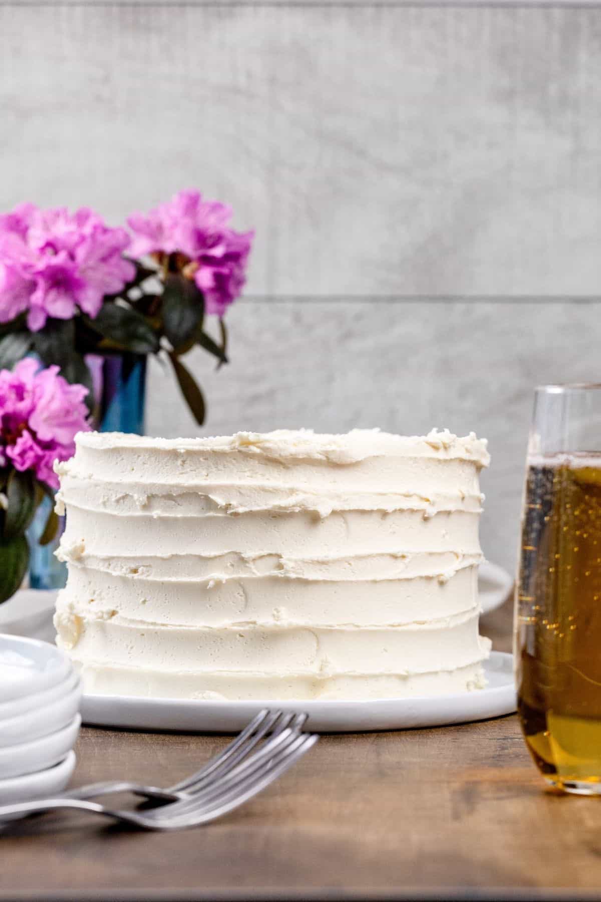 A vegan vanilla cake covered in vanilla frosting is on a round white plate on the kitchen table. It's next to a stack of plates, forks, and a glass of champagne. Pink flowers are blurred in the background.