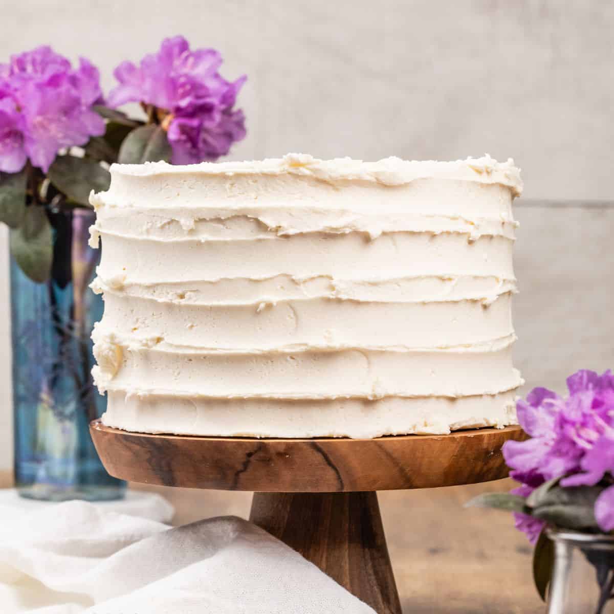 A vegan vanilla cake covered in vanilla frosting is on a wood cake stand. Vases of small purple flowers are next to the cake.