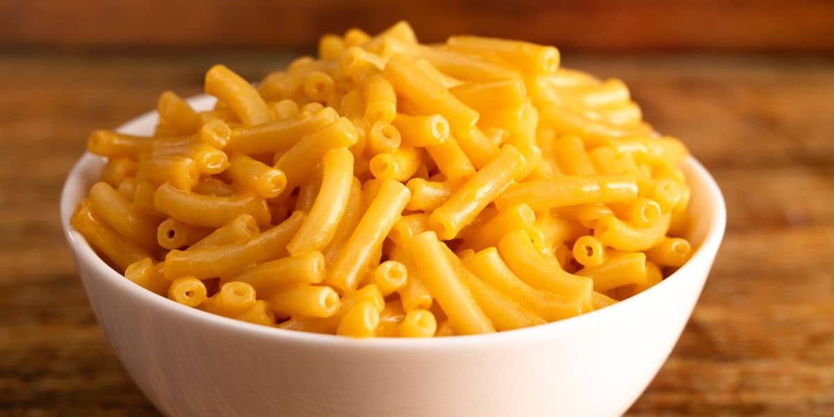 A white bowl is filled with Mac and cheese. It rests on a wood tabletop.