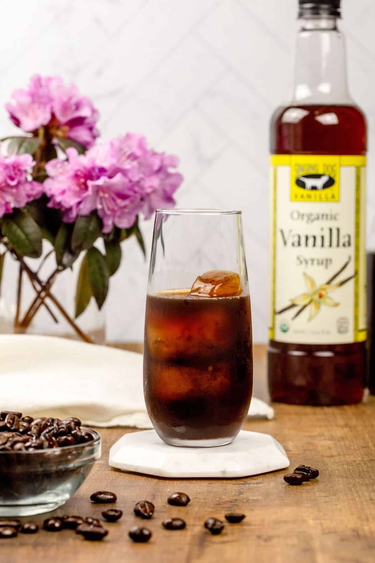 A glass filled with coffee ice cubes and cold brew coffee on a wood tabletop. A jar of vanilla syrup and flowers are blurred in the background. Coffee beans are in front of the cup of coffee.