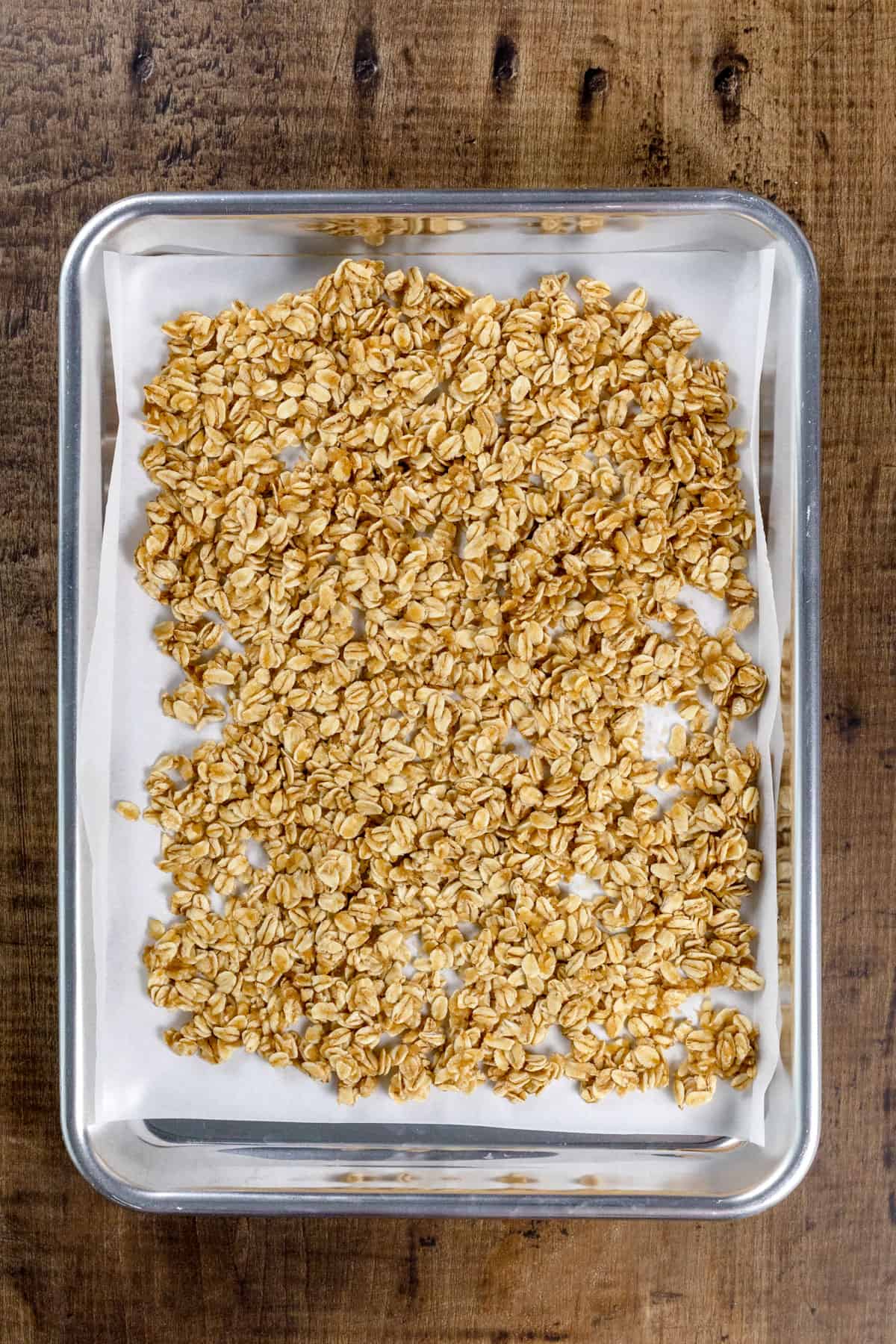 Baking sheet covered in cooked granola on a white parchment paper on a wood kitchen table.