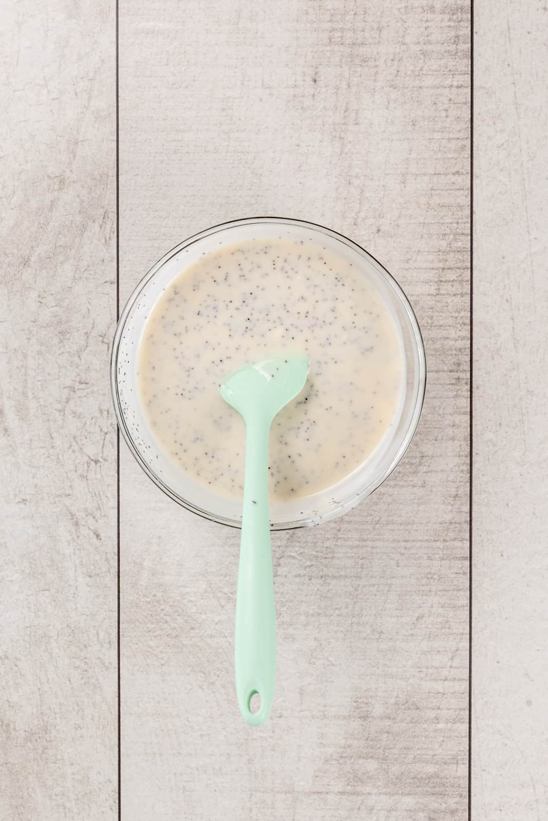 A glass bowl on a white wood tabletop is filled with dairy free poppyseed dressing. A light green spatula is in the dressing.