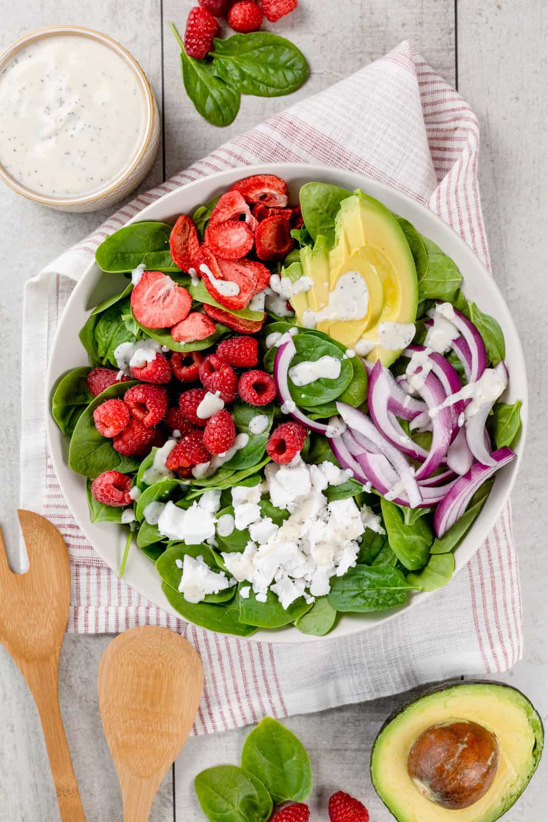 A big white bowl filled with raspberry and strawberry poppyseed salad. It is on a white table with a red and white napkin under the bowl. A bowl of dressing and more ingredients surround the bowl.