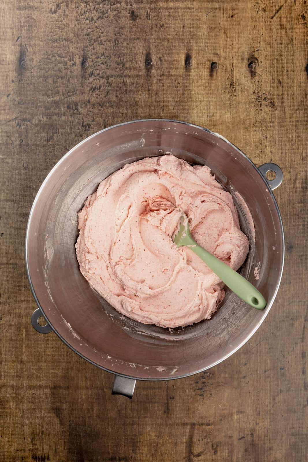 A silver mixing bowl is on a wood tabletop filled with finished buttercream frosting. A green spatula is in the frosting.