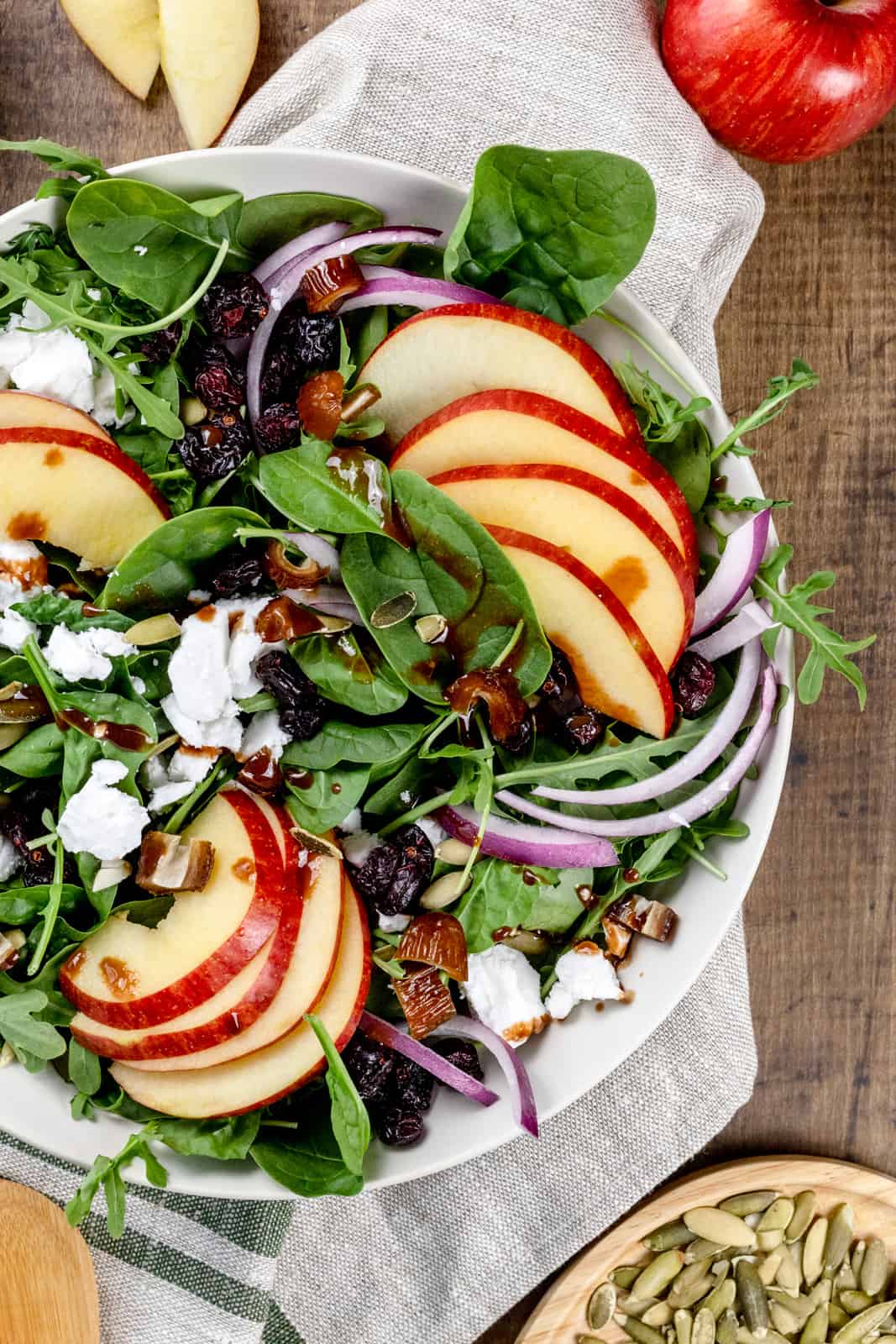 Closeup of a big bowl of a fuji apple salad filled with greens, apples, red onion, and other ingredients. A towel and other ingredients surround the bowl.