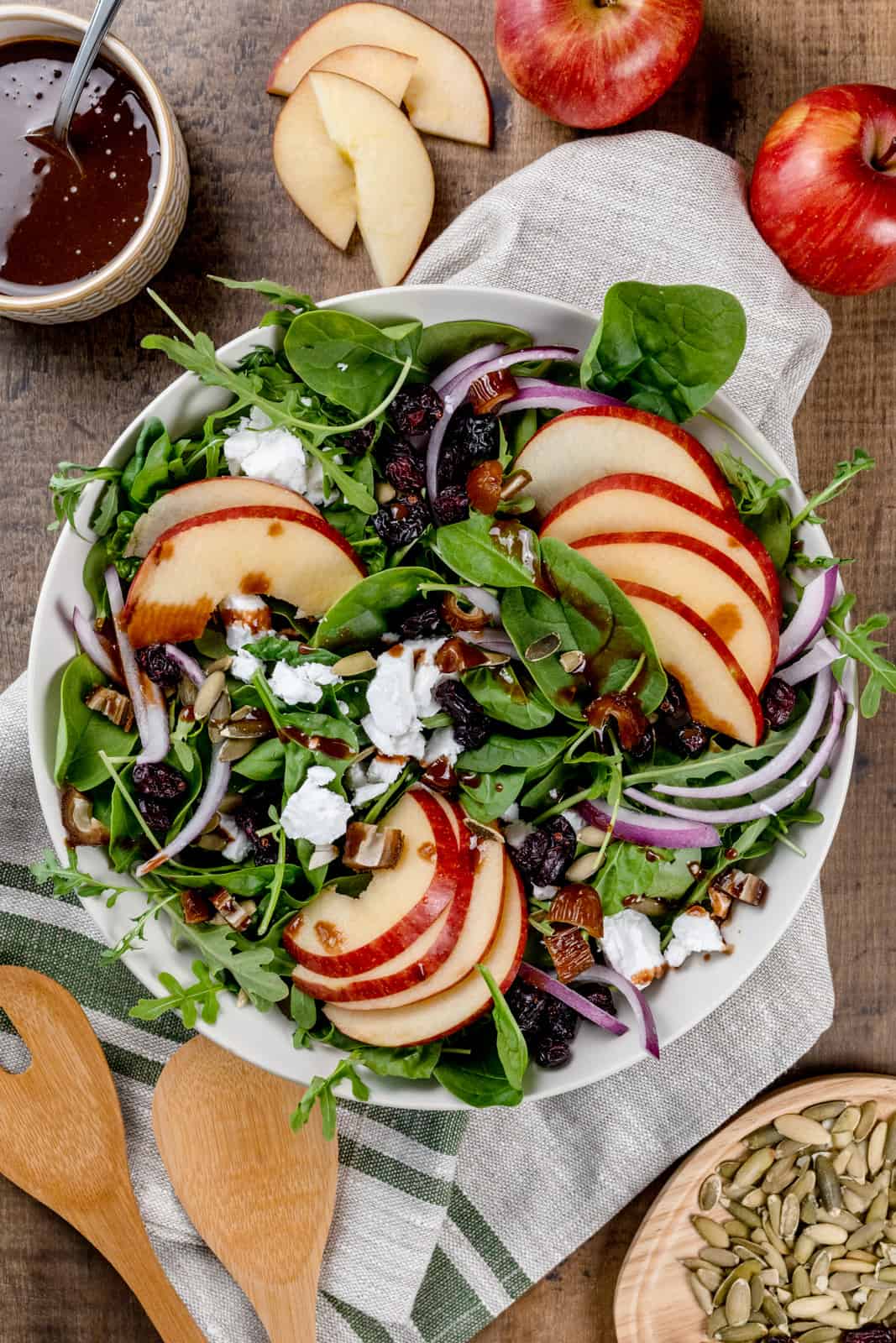 A big bowl of a salad filled with apples, greens, red onion, feta cheese, and dried fruits. It's on a wood tabletop surround by other ingredients like apples and pumpkin seeds. A small bowl of extra balsamic dressing is in the top left.