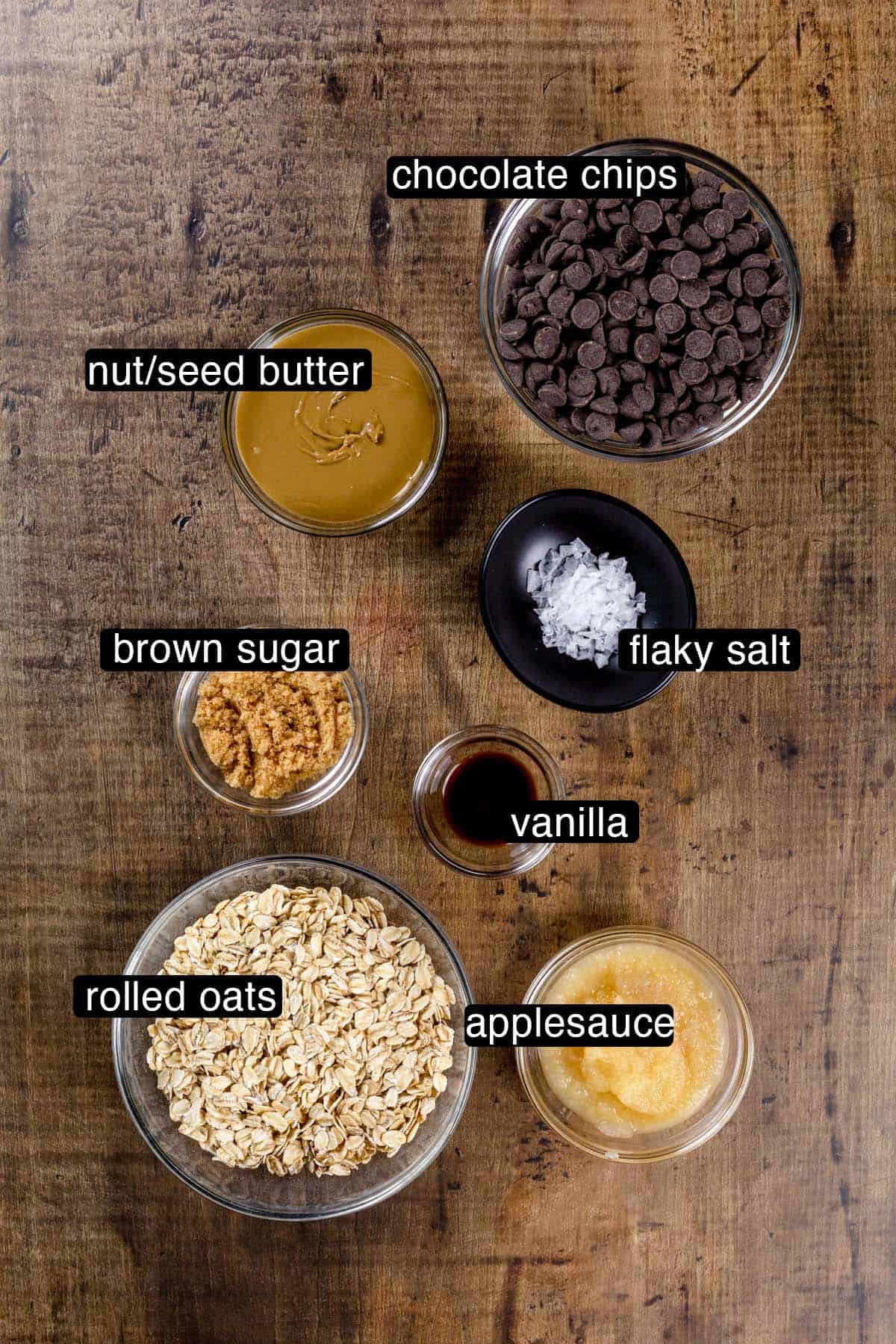 Ingredients for granola bark in various glass bowls on a wood table top. Black and white labels have been added to name each ingredient like oatmeal and applesauce.