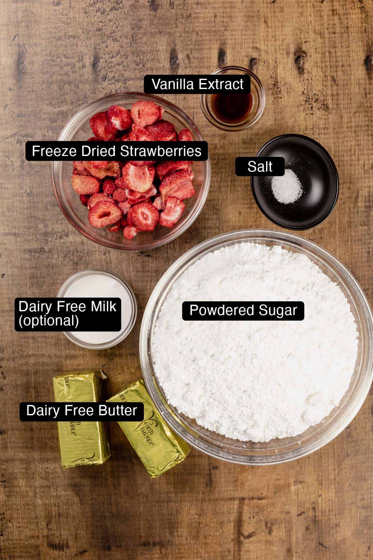 Ingredients for vegan strawberry frosting in various glass bowls on a wood tabletop. Black and white labels have been added to name each ingredient.