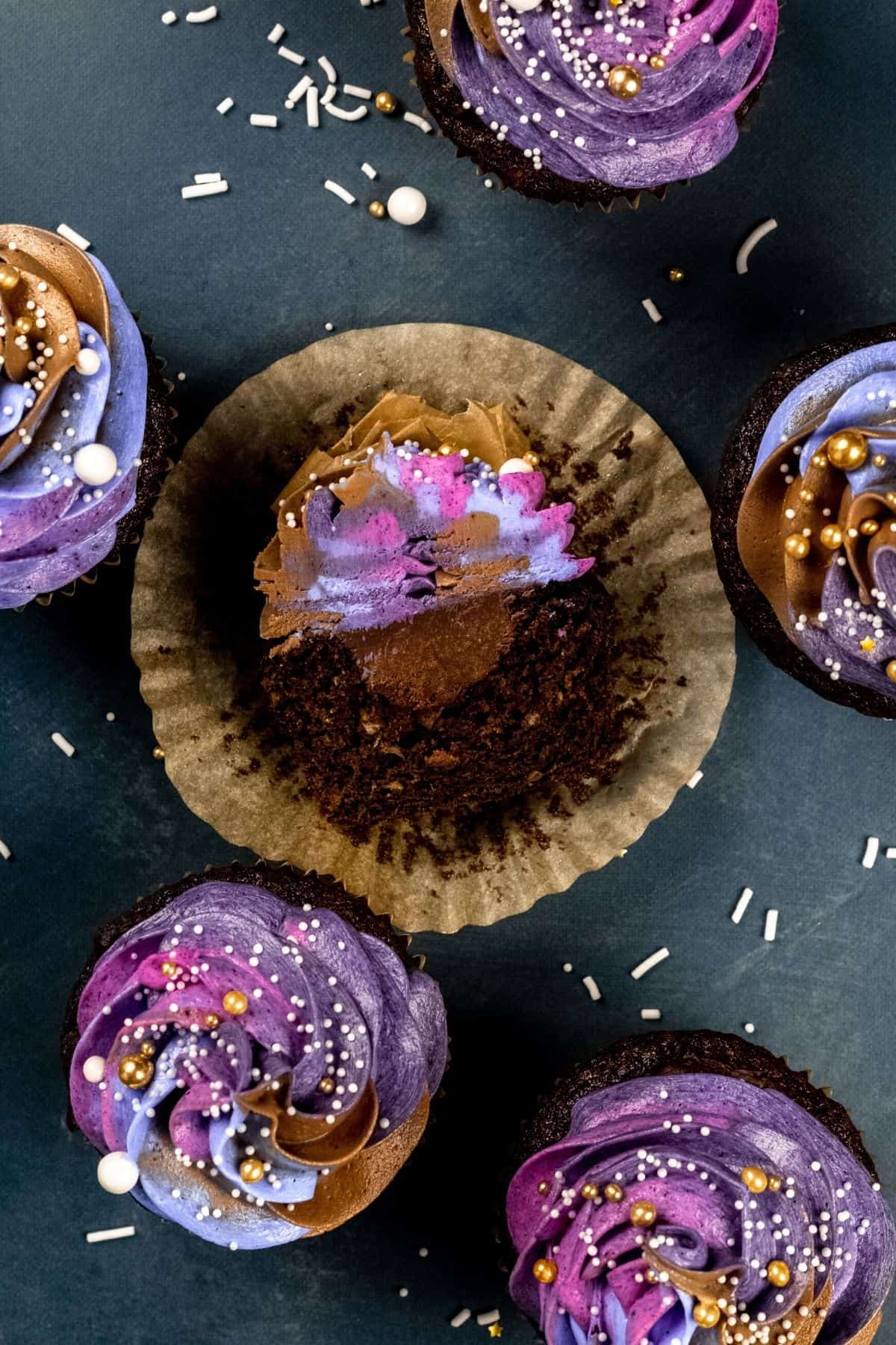 Looking down at a chocolate cupcake filled with chocolate ganache and lots of colorful swirled frosting on top. Sprinkles and other cupcakes surround this one that is cut in half and laying down on its side.