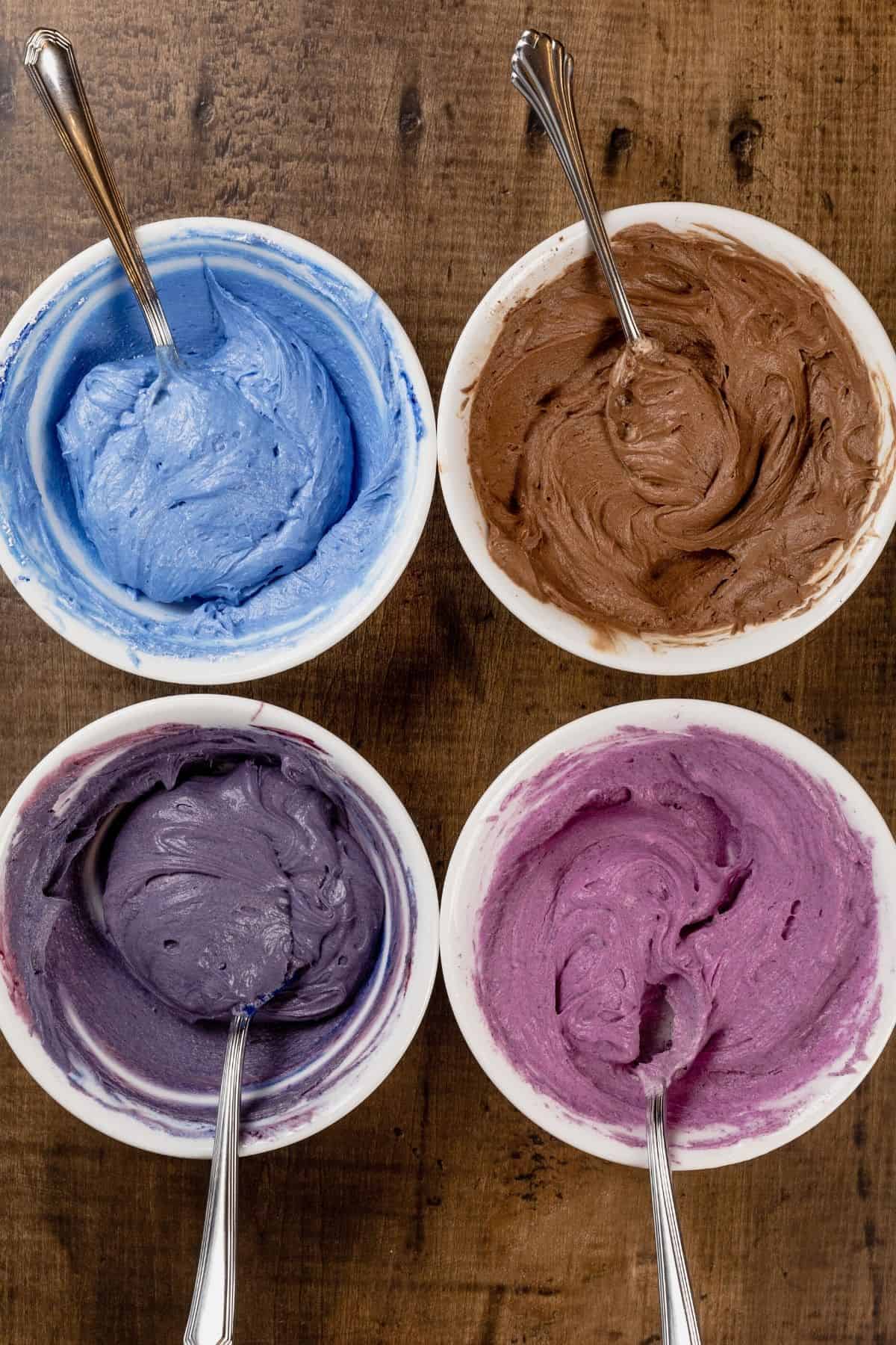 4 white bowls filled with different colors of frosting on a wood tabletop with spoons in each of them.