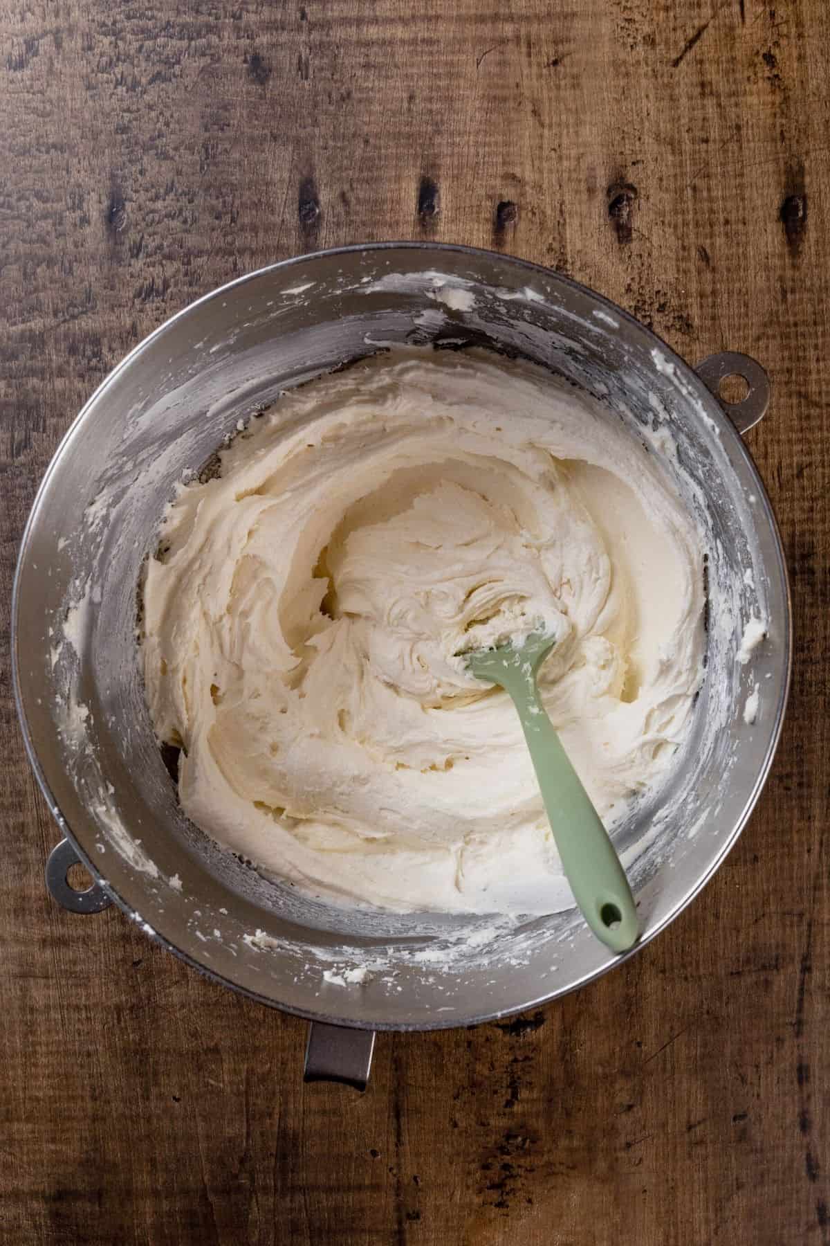A silver mixing bowl is filled with finished vegan frosting. It sits on a wood table. A green spatula is in the frosting.