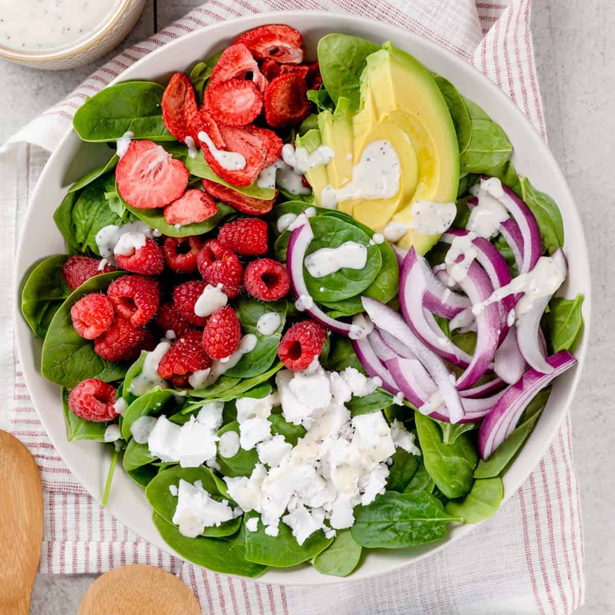 Big bowl filled with a raspberry and strawberry spinach salad with dairy free poppyseed dressing. Other ingredients like red onion and dairy free feta cheese and avocado are also on the salad.