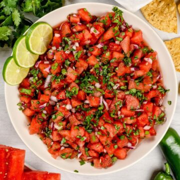 Bowl filled with tomato free salsa and lime slices. A bunch of chips, fresh cilantro, watermelon slices, and peppers surround the bowl.