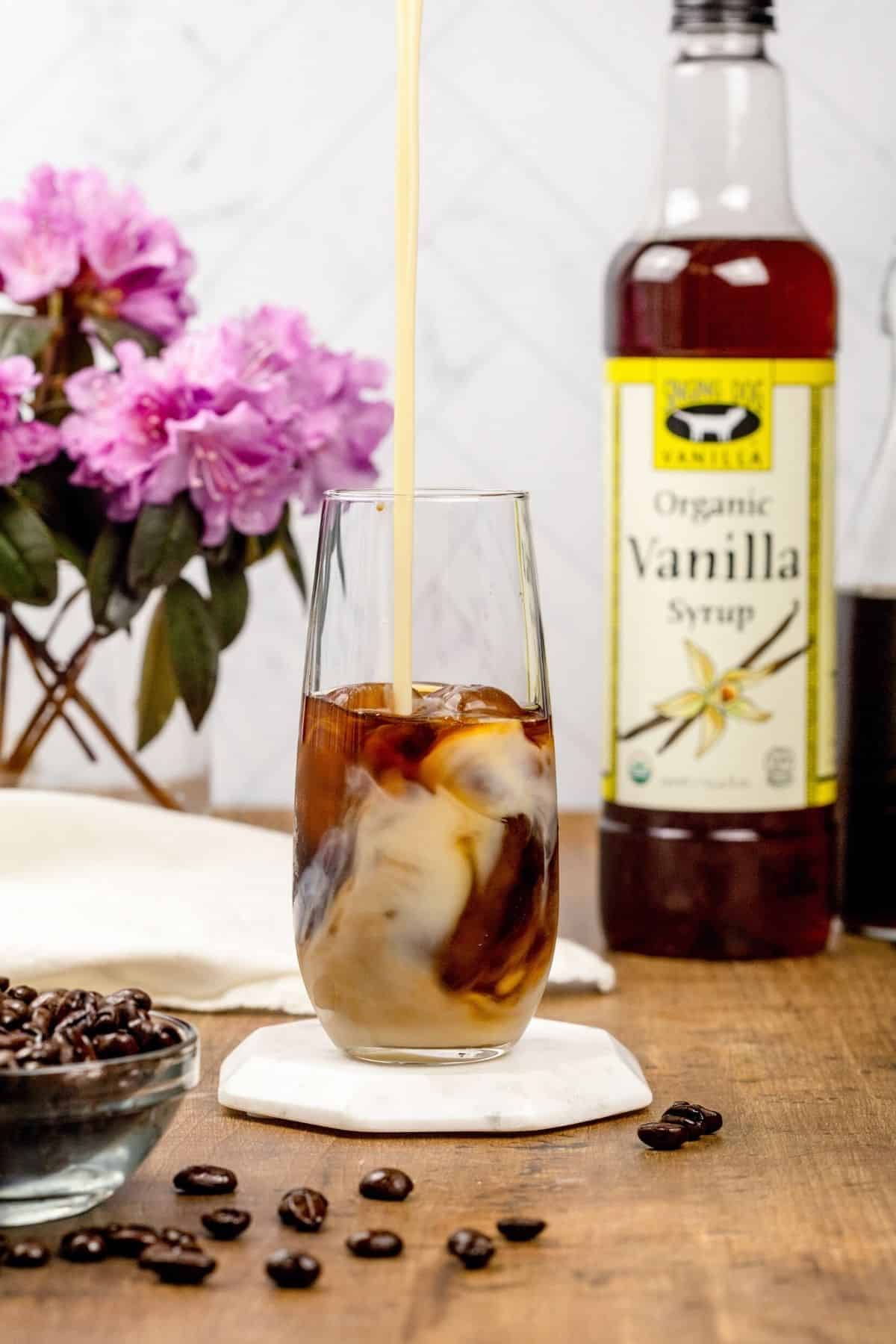 Pouring sweet vanilla cream into a glass cup on a wood tabletop. A jar of vanilla syrup and flowers are blurred in the background. Coffee beans are in the front of the cup.