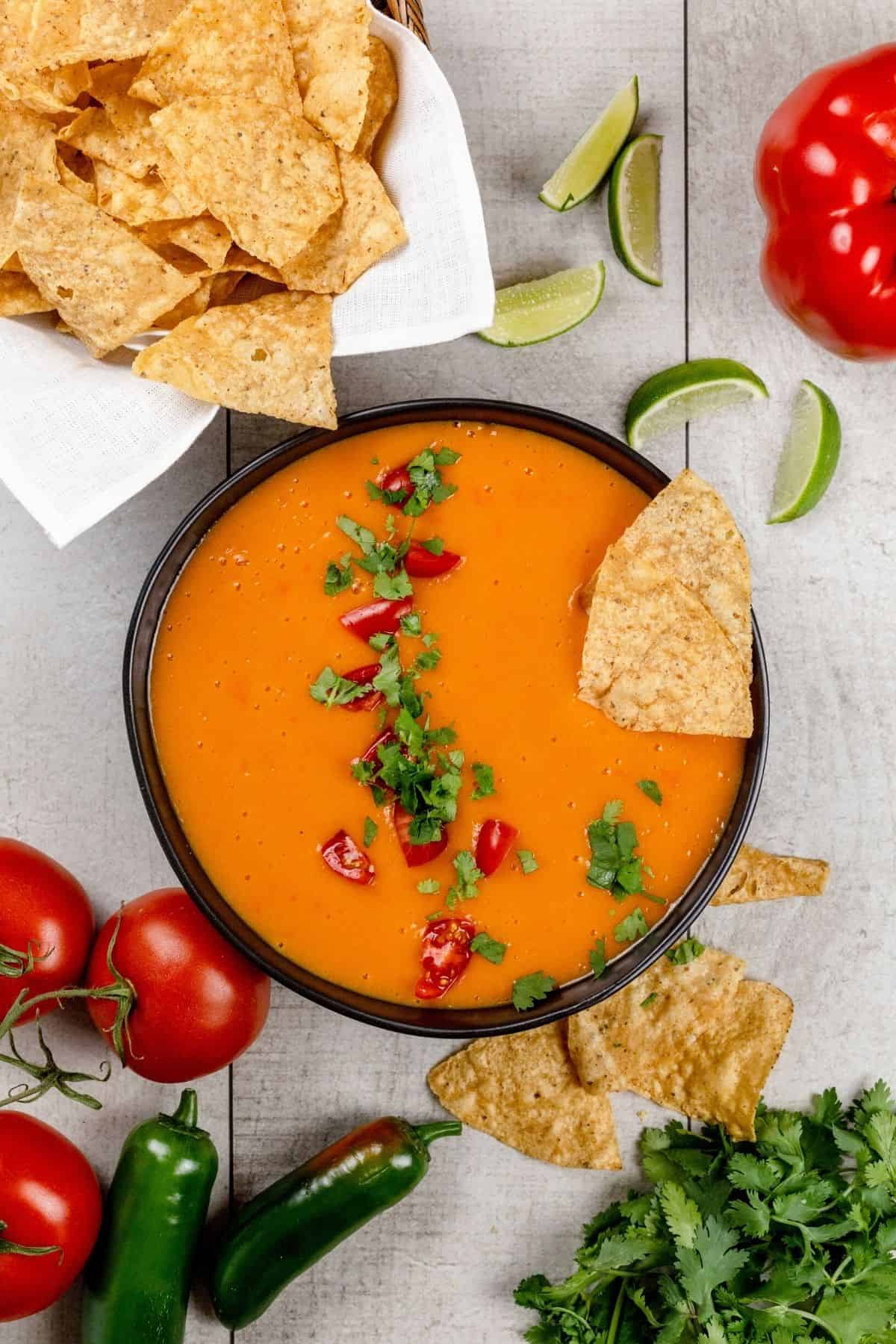 A big bowl filled with vegan cheese sauce is on a white tabletop. It's topped with cilantro and tomatoes. A few chips are sticking in the sauce. More chips in a bowl are next to the sauce. It's surrounded by fresh ingredients.