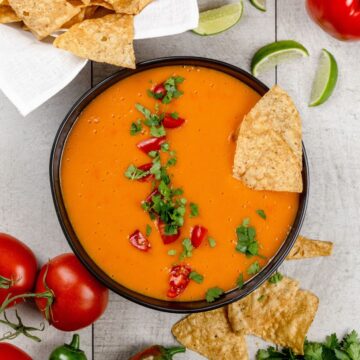 A black bowl is filled with vegan cheese sauce. Tiny tomatoes and fresh cilantro are scattered on top of the sauce. Some chips are dipped into the sauce. More ingredients, tomatoes, and chips are next to the bowl.