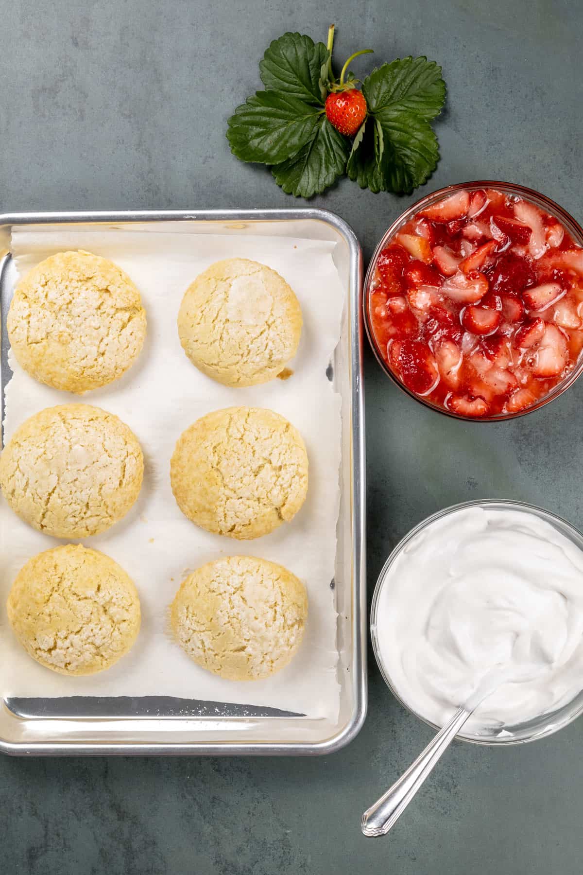 A tray filled with the cooked and cooled shortcakes, a bowl filled with the strawberries, and a bowl filled with dairy free whipped cream on a countertop.